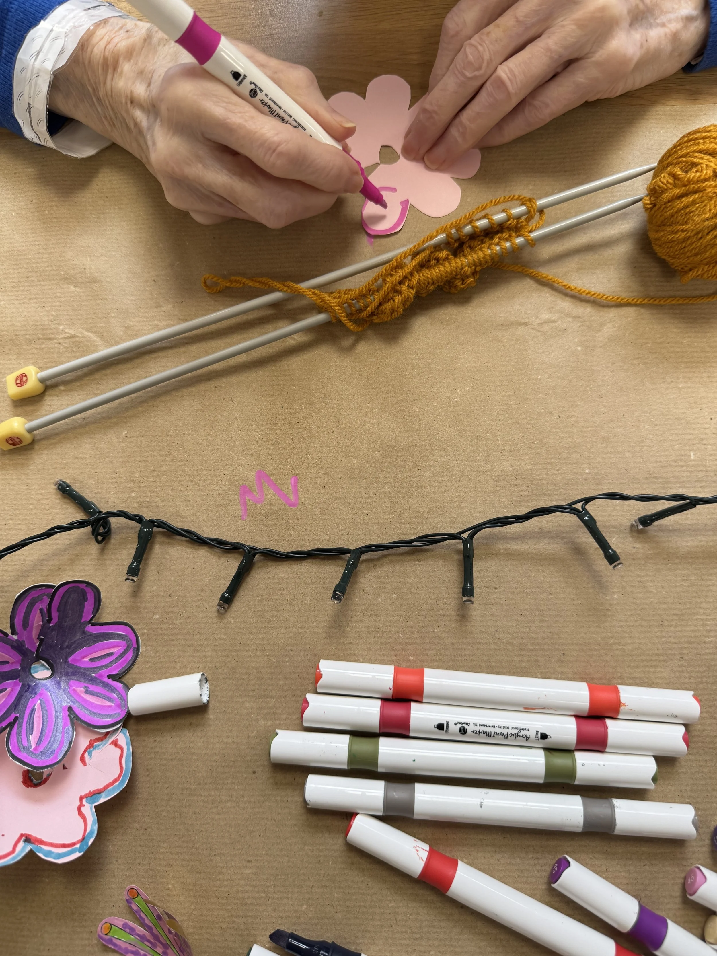 Person drawing a pink paper flower with a purple marker, with knitting needles, yellow yarn, and colorful pens on a brown table.