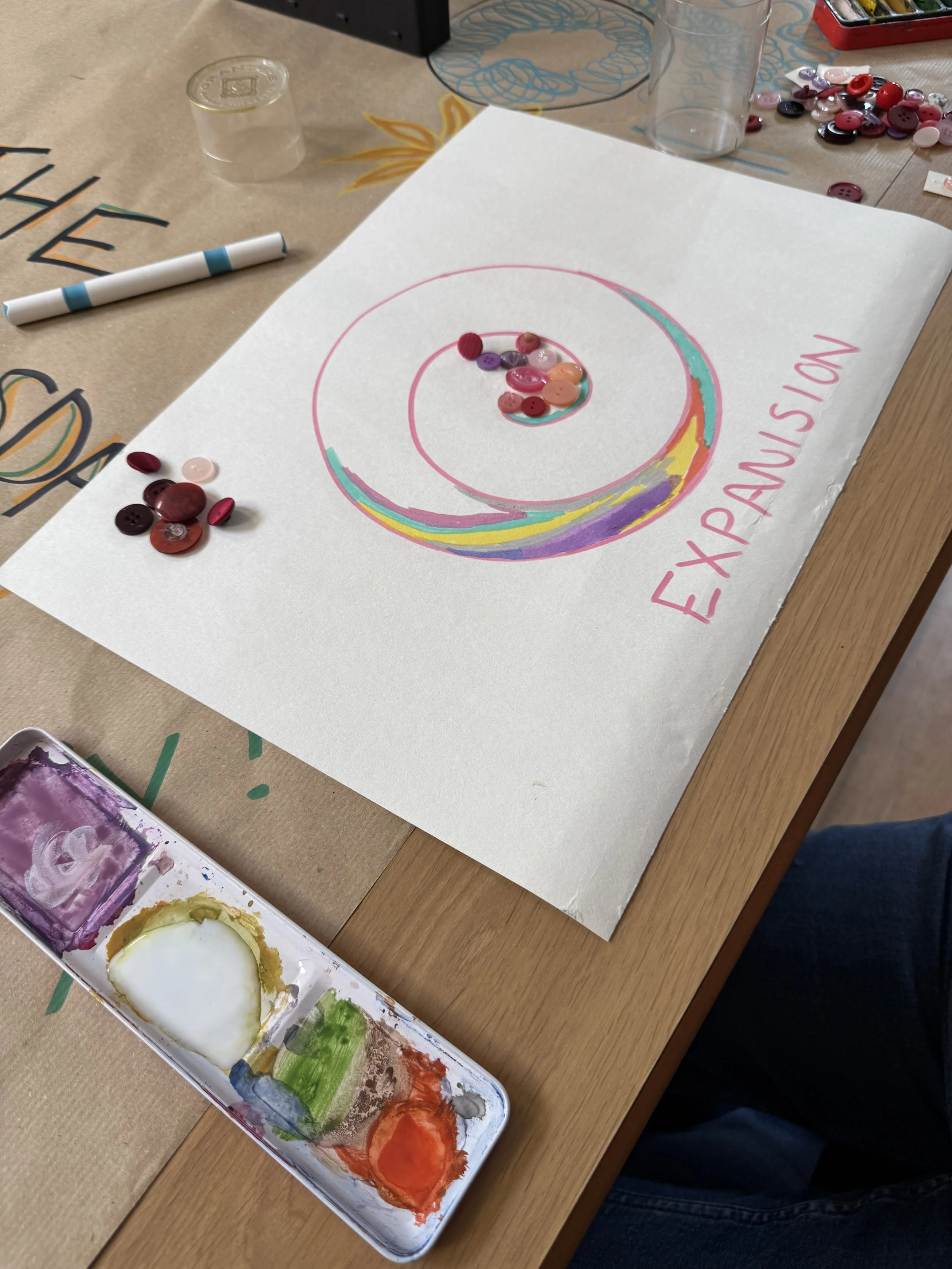 A painted poster with colorful concentric circles and the words 'EXPAUS ION' written on the side, surrounded by buttons, paint palette, and art supplies.