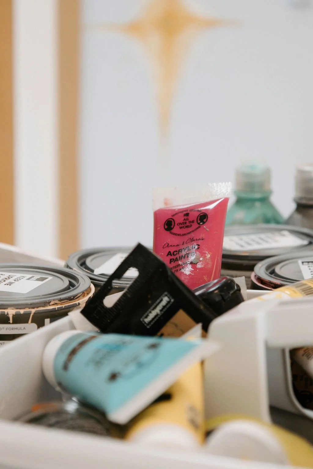 A collection of various paint containers and tubes of paint on a table, including cans of paint and small tubes of acrylic paint, with a blurred background.