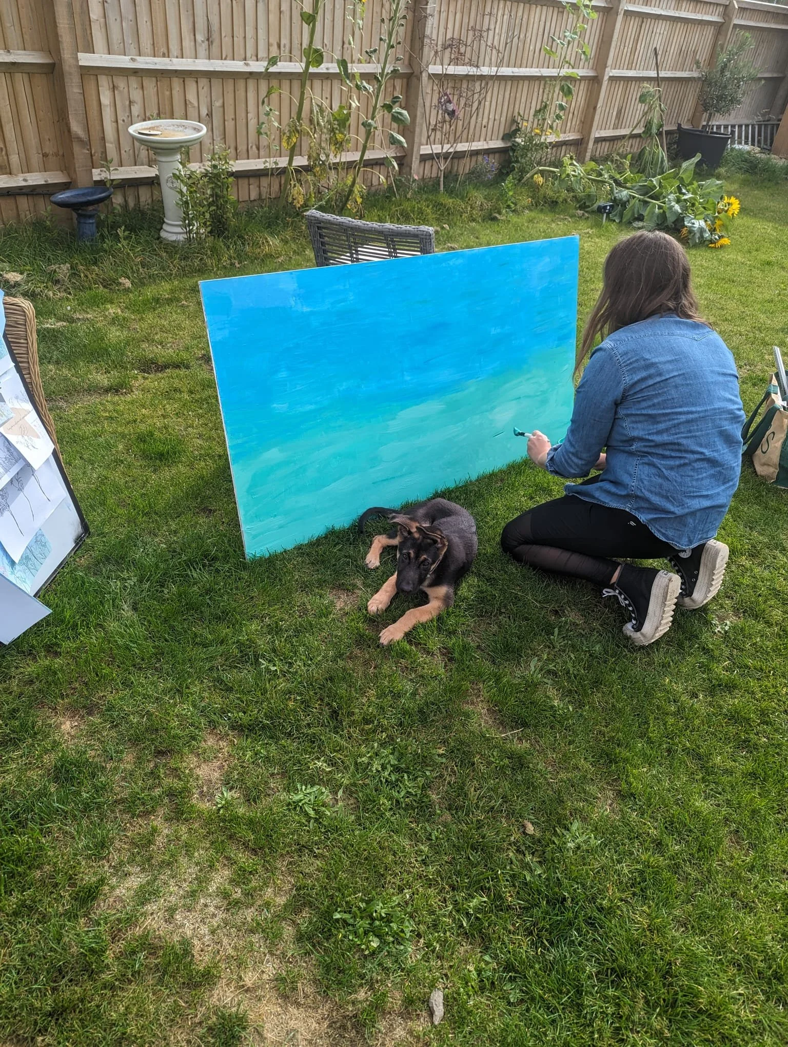 A woman painting a large blue canvas outdoors in a grassy backyard with a dog lying nearby.