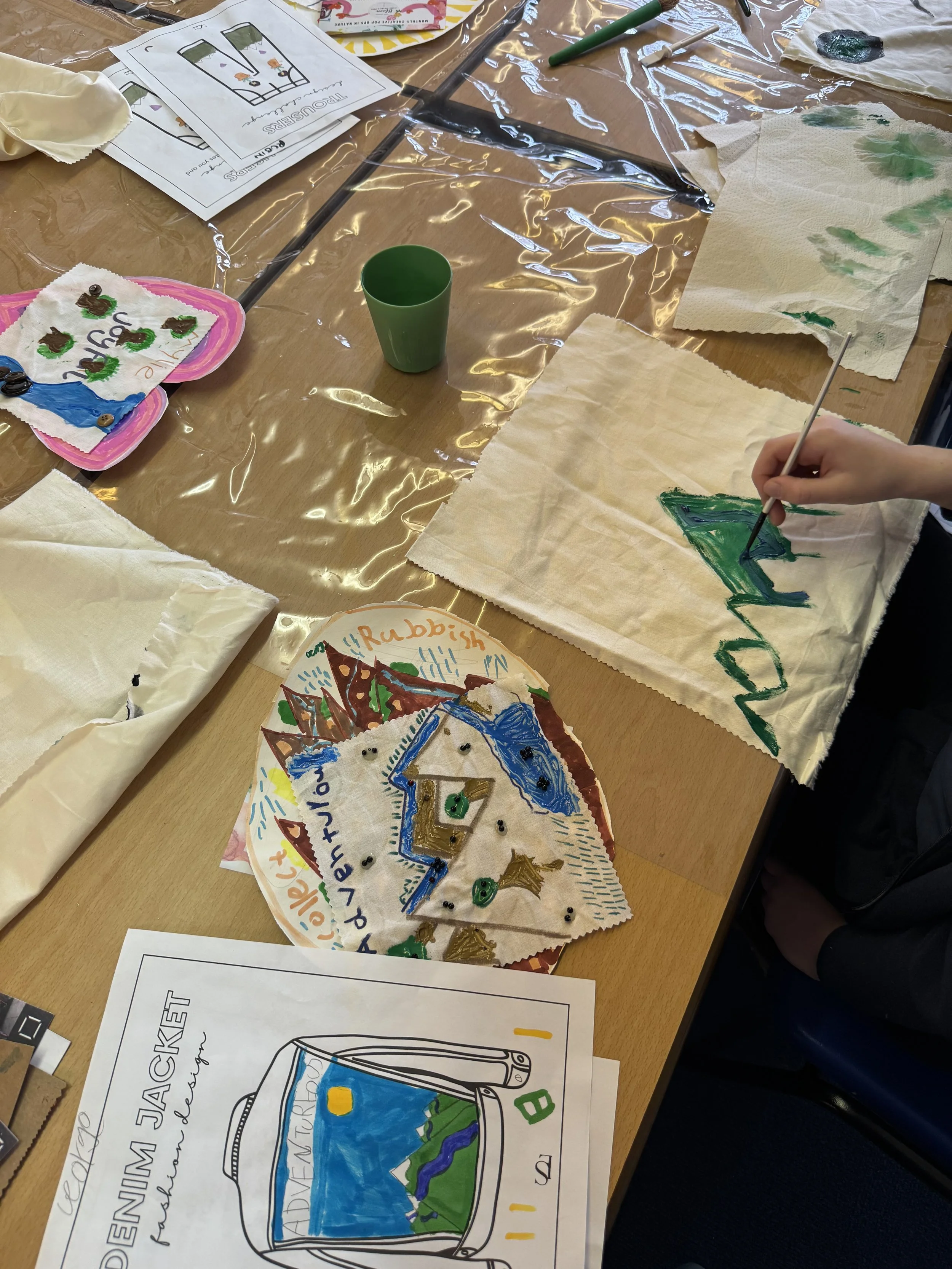 A classroom craft activity with papers, paints, and drawing materials on a table. There are decorated paper plates with drawings and words, some papers with landscape drawings, and instructions for creating a travel journal.