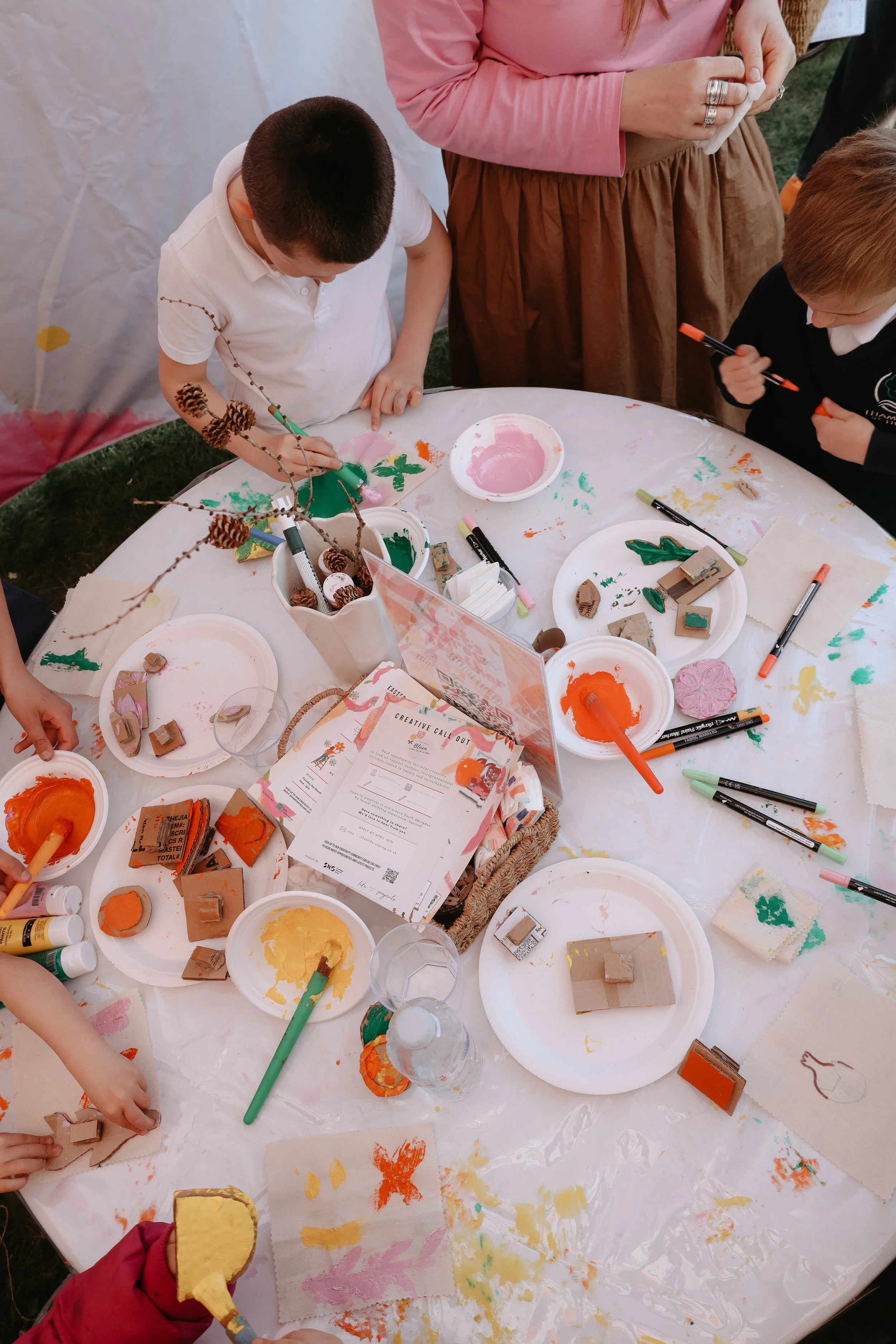Children at a craft table painting and stamping with various colors and stamps during an outdoor event.