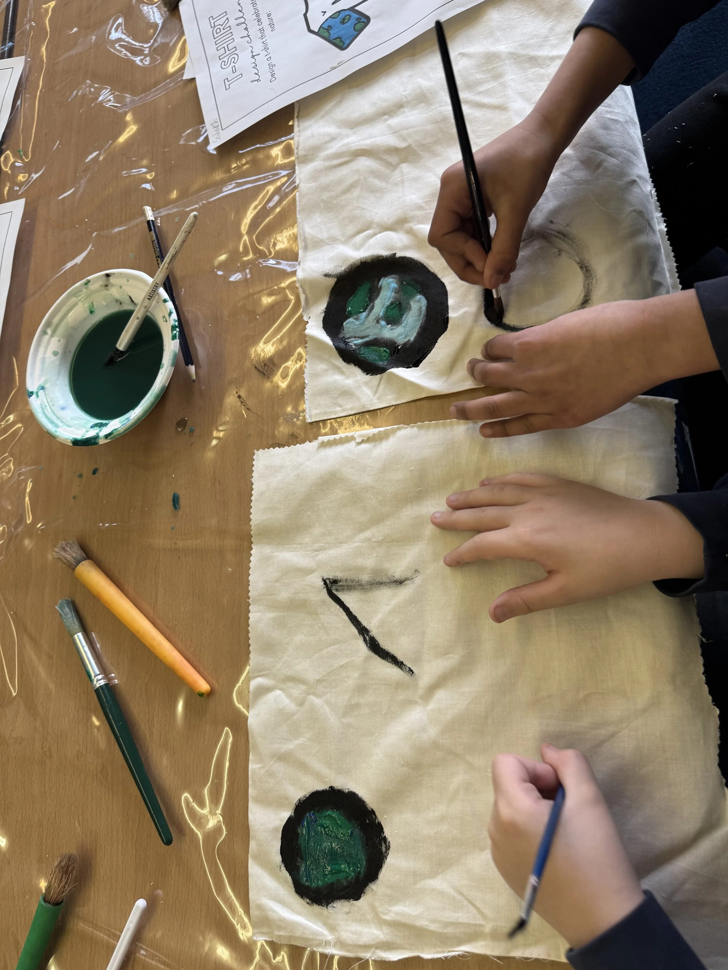 Children are painting and drawing on large sheets of paper with brushes and black paint, creating circular and zigzag shapes on a wooden table.