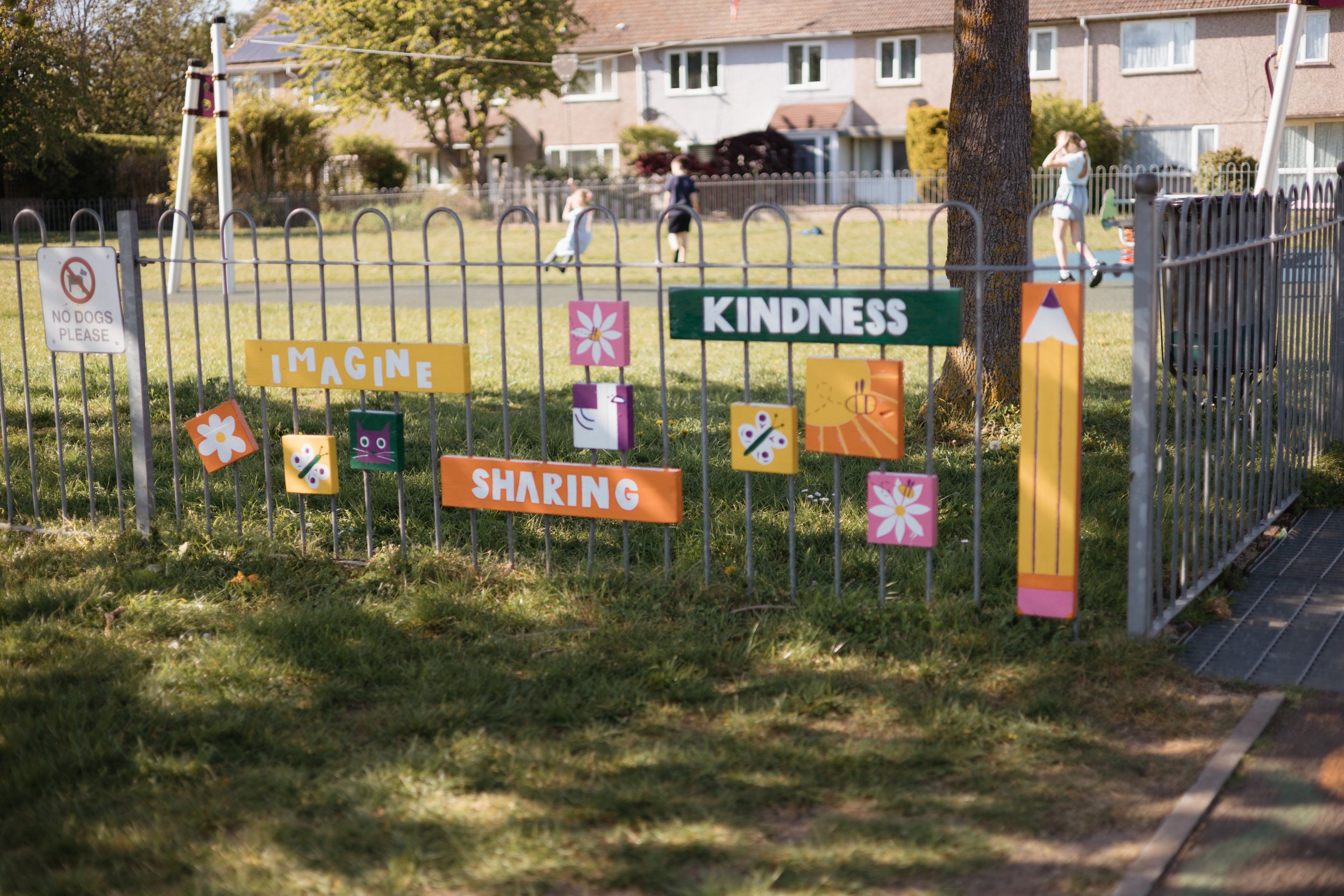 A fenced playground with colorful signs that read 'IMAGINE,' 'KINDNESS,' and 'SHARING.' Children are playing on the grass in the background, and there are trees and houses nearby.