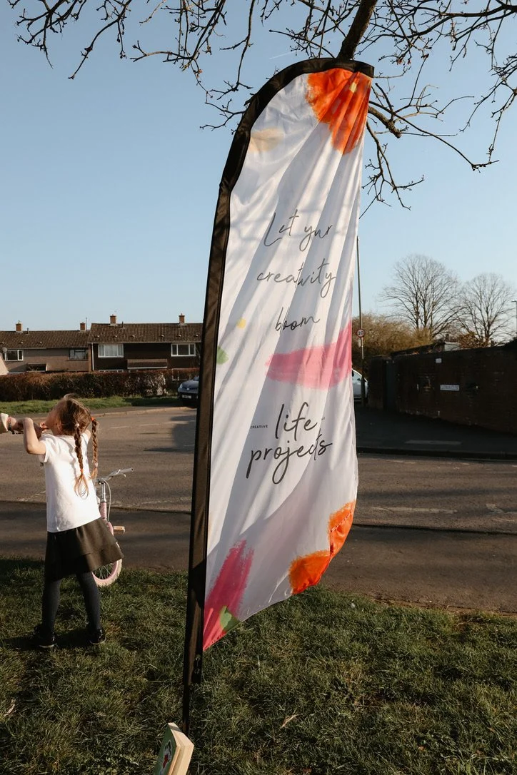 A colorful outdoor banner with the text 'Let your creativity bloom' and 'life projects,' standing on a grassy area next to a sidewalk. A young girl with braided hair, wearing a white top and dark skirt, is playing with a pink bicycle nearby. In the background, there are houses, leafless trees, and parked cars.