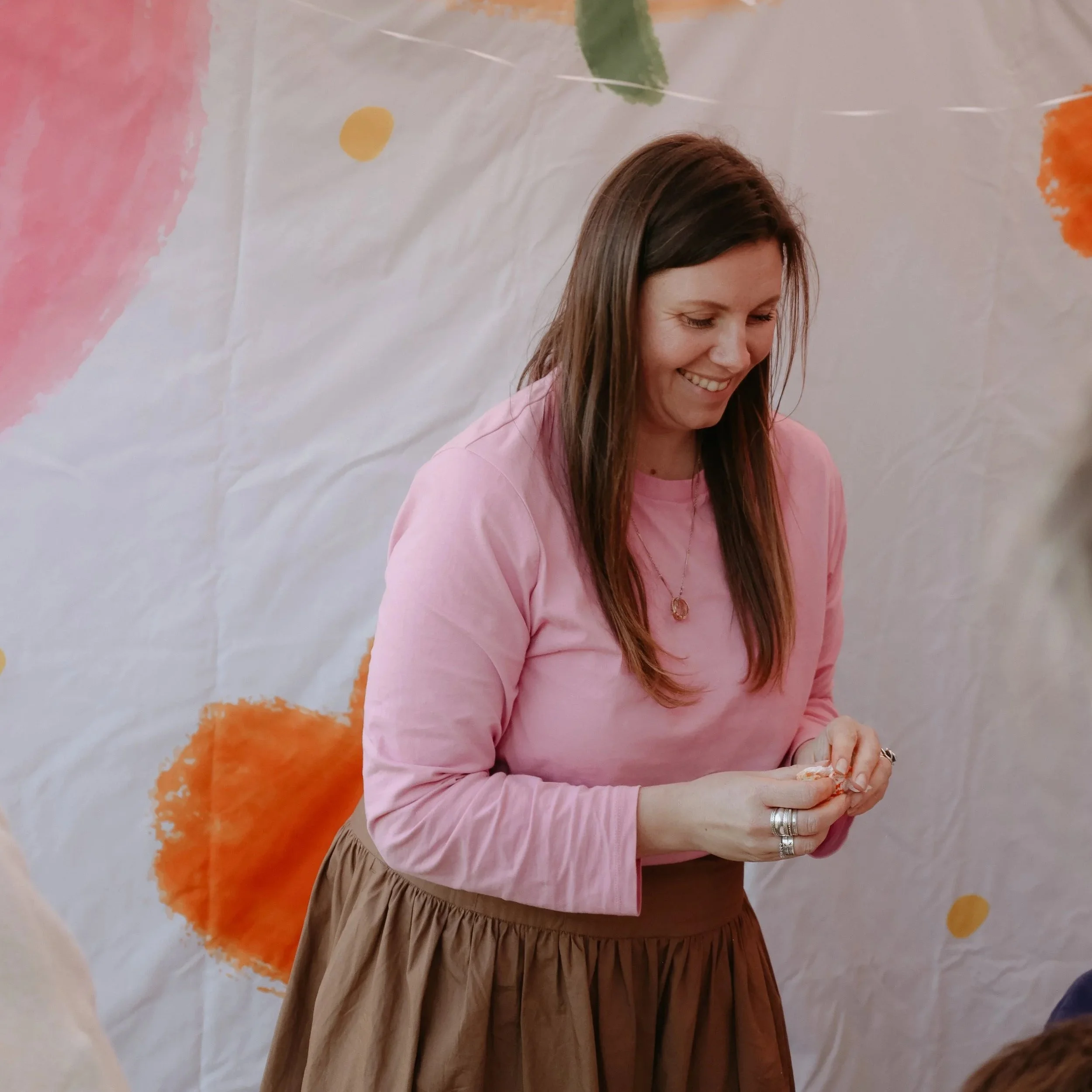 A smiling woman with long brown hair wearing a pink long-sleeve shirt and a brown skirt, standing in front of a white backdrop with pink, orange, and yellow abstract shapes.