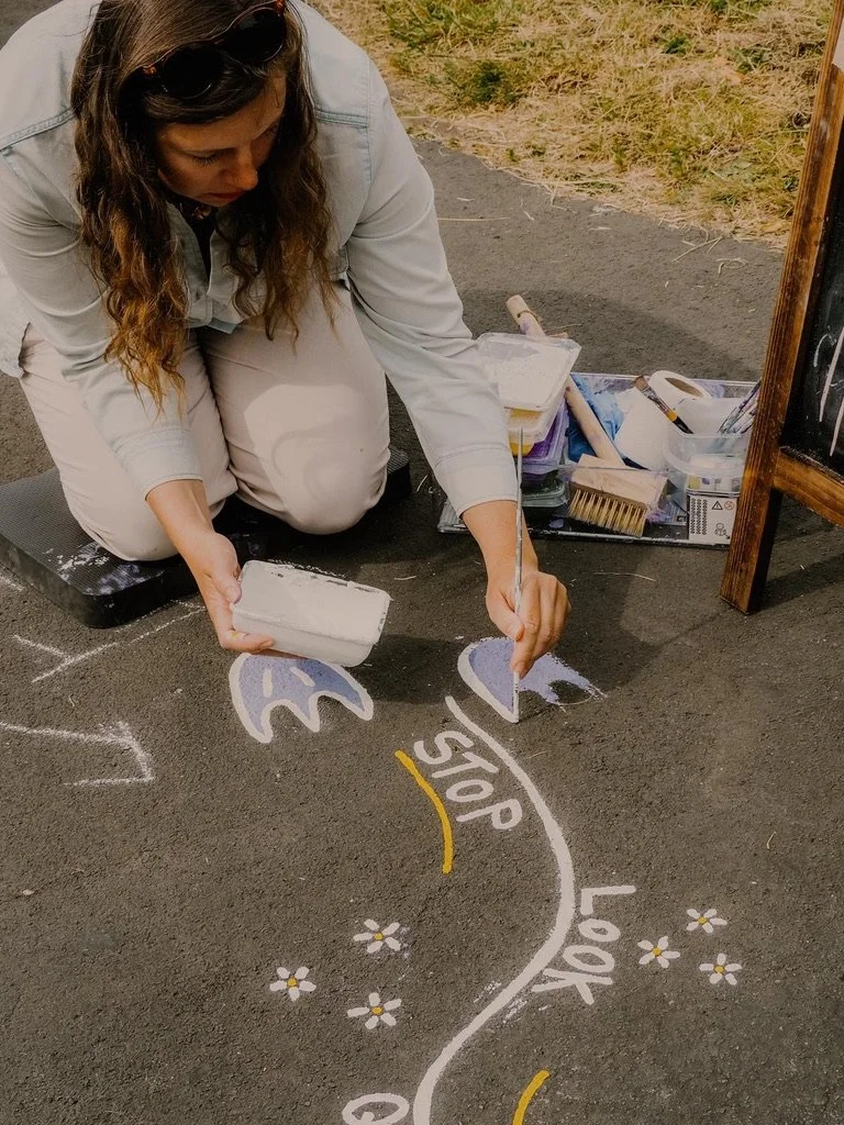 Woman drawing a classroom crossing and stop sign on the pavement with chalk, outdoors, with art supplies nearby.