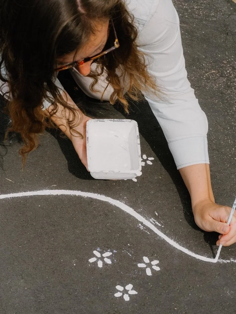 A woman wearing glasses and a white shirt is drawing white flower shapes and a wavy line on a black asphalt surface using white paint and a small brush.