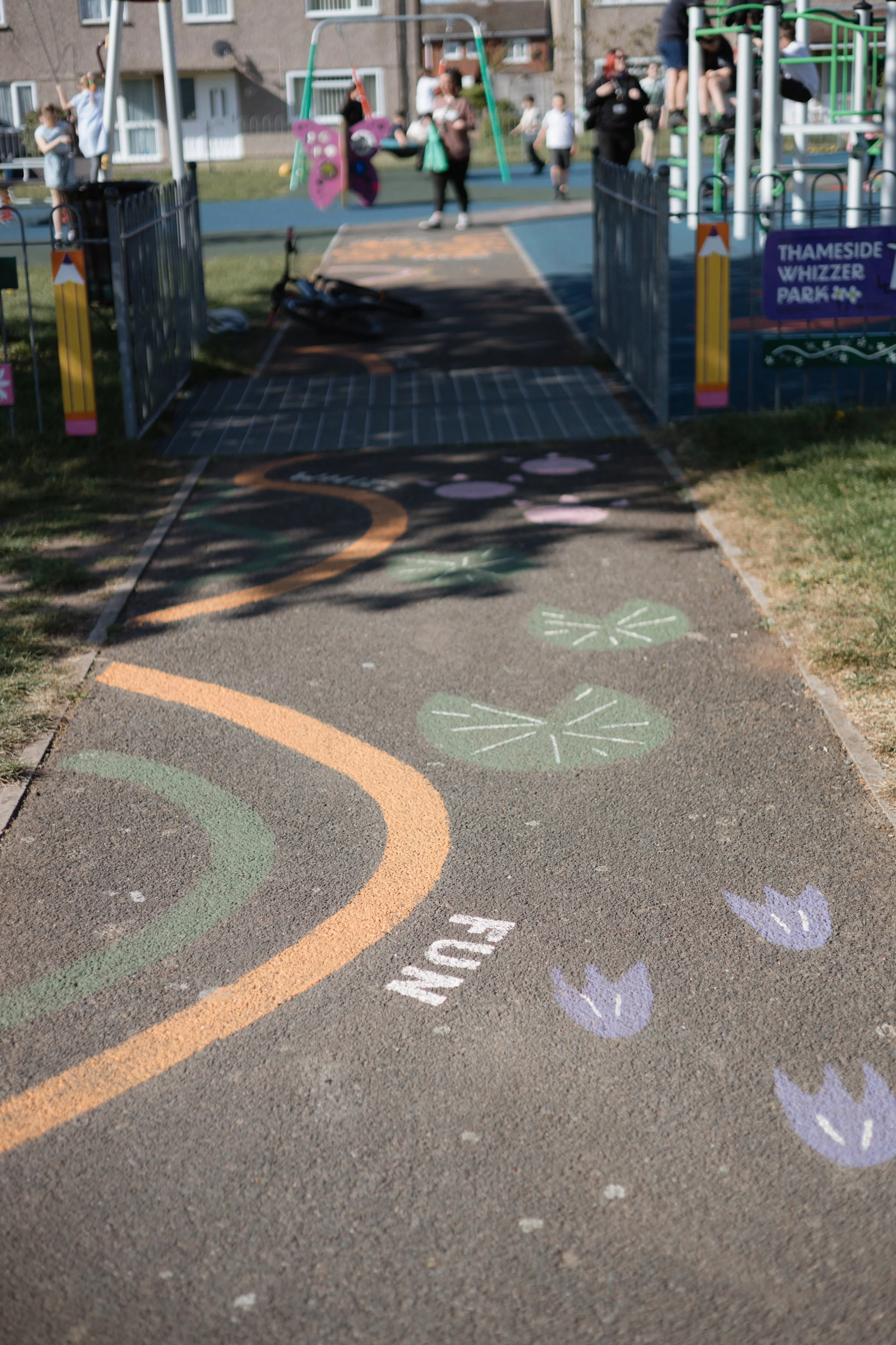 A playground with a walking path decorated with colorful chalk drawings, including flowers, leaves, and a winding line. In the background, there are children and adults near the playground equipment, which includes swings and climbing structures. The area is fenced, and there are residential buildings nearby.