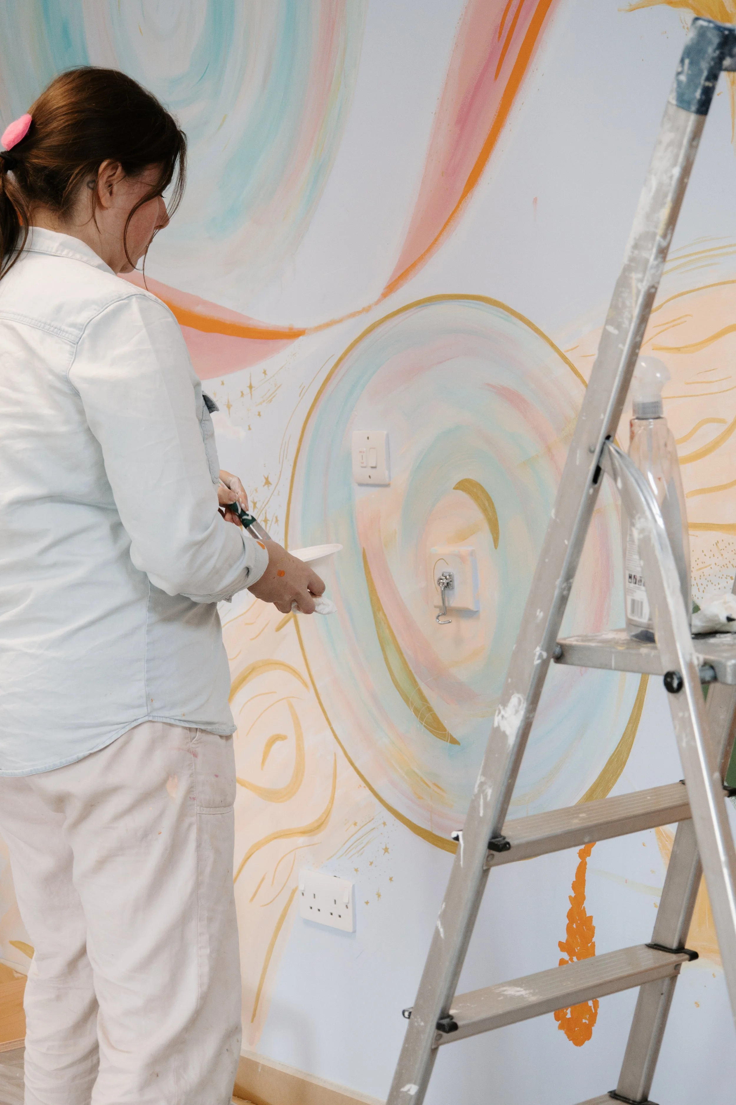 A woman painting a colorful wall mural with pastel swirls and gold accents. She is wearing a white shirt and beige pants, standing next to a silver ladder.