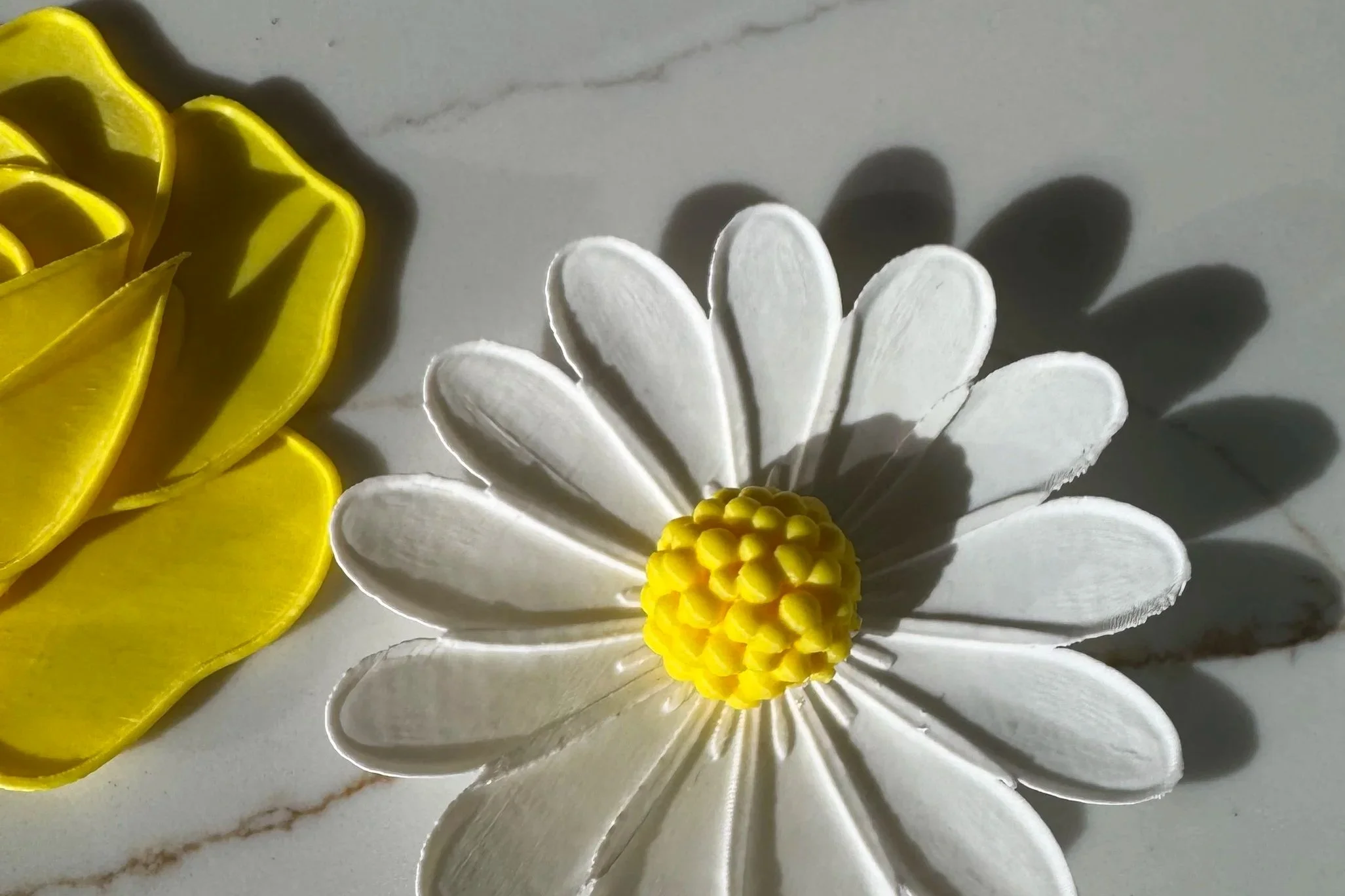 A close-up of a decorative ceramic or plastic white daisy with a yellow center, next to a yellow flower-shaped object, both casting shadows on a gray marble surface.