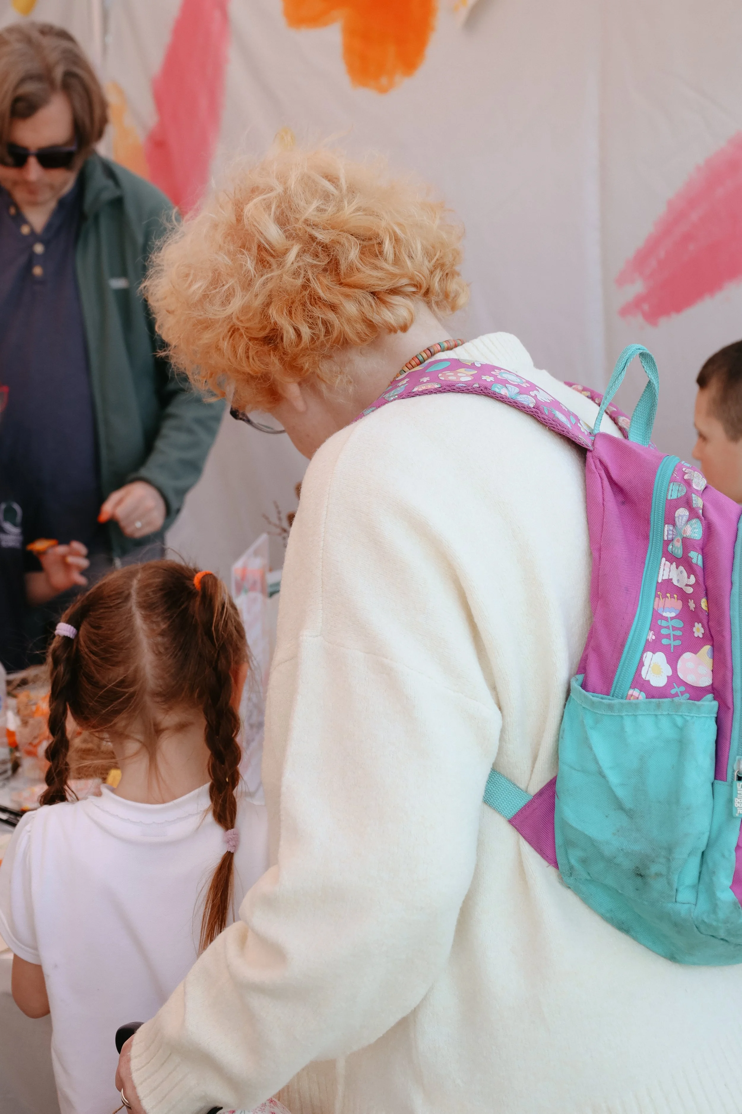 A woman with curly red hair and a colorful backpack leaning towards a young girl with braided hair in a white shirt at an indoor event with pink and orange decorations.