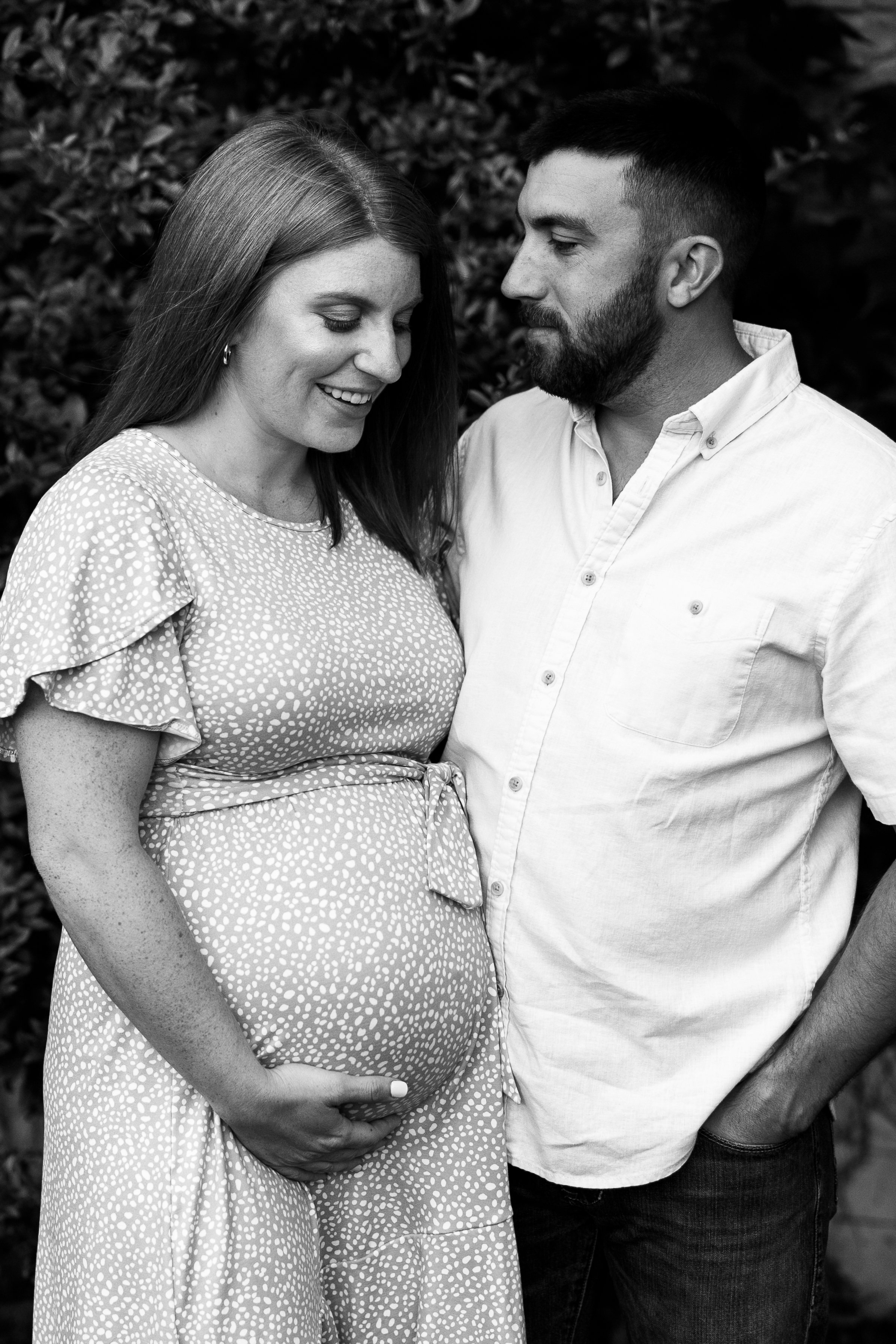 Black and white photo of a pregnant woman and a man, likely her partner, standing close together outdoors, smiling and looking at each other. The woman is wearing a polka-dot dress with flutter sleeves, and the man is in a light-colored button-up shi