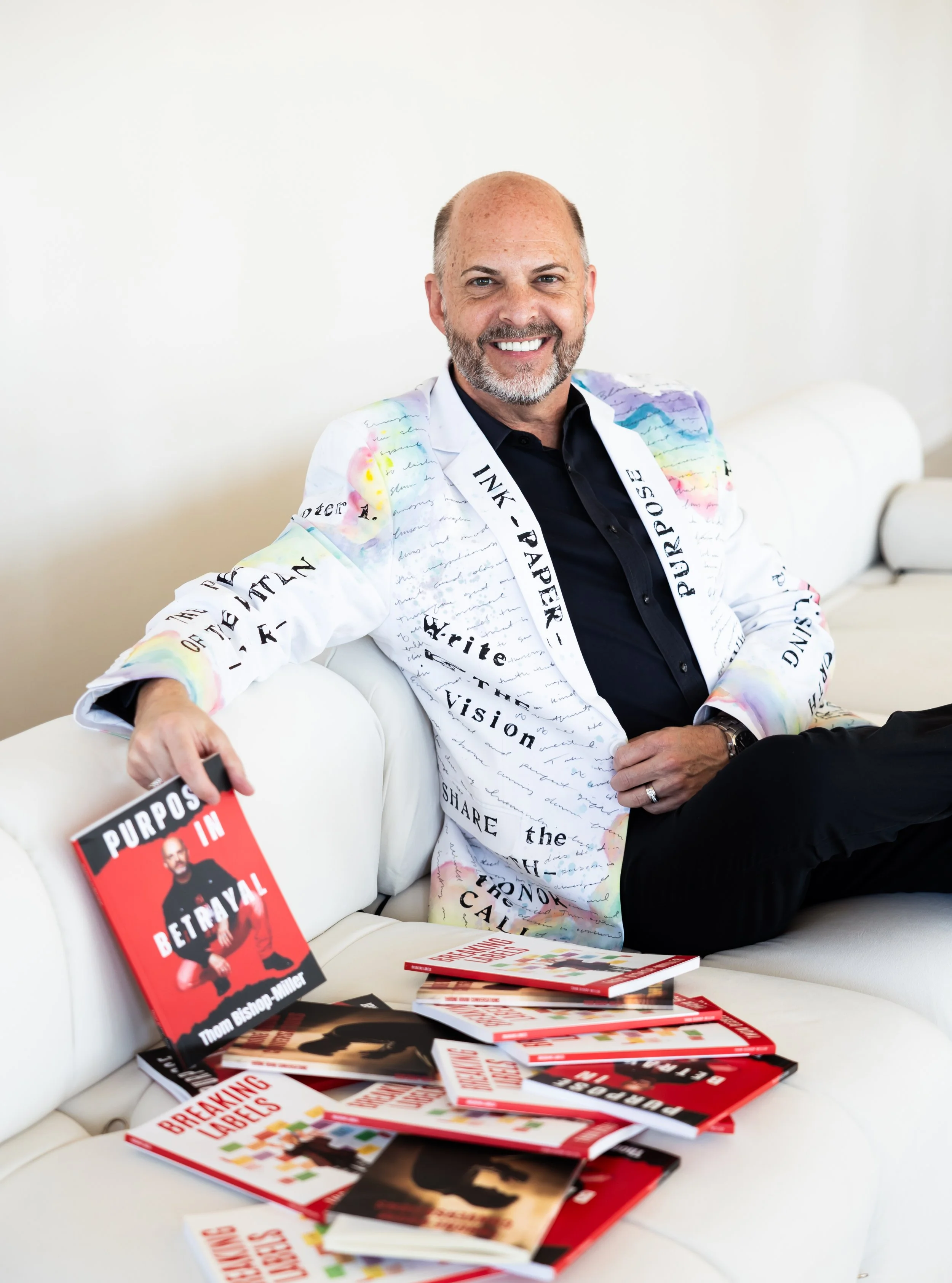 A man with a beard smiling at the camera, sitting on a white couch, wearing a white jacket with colorful writing and a black shirt, surrounded by books and a book titled 'Purposes in Betrayal' in his hand.