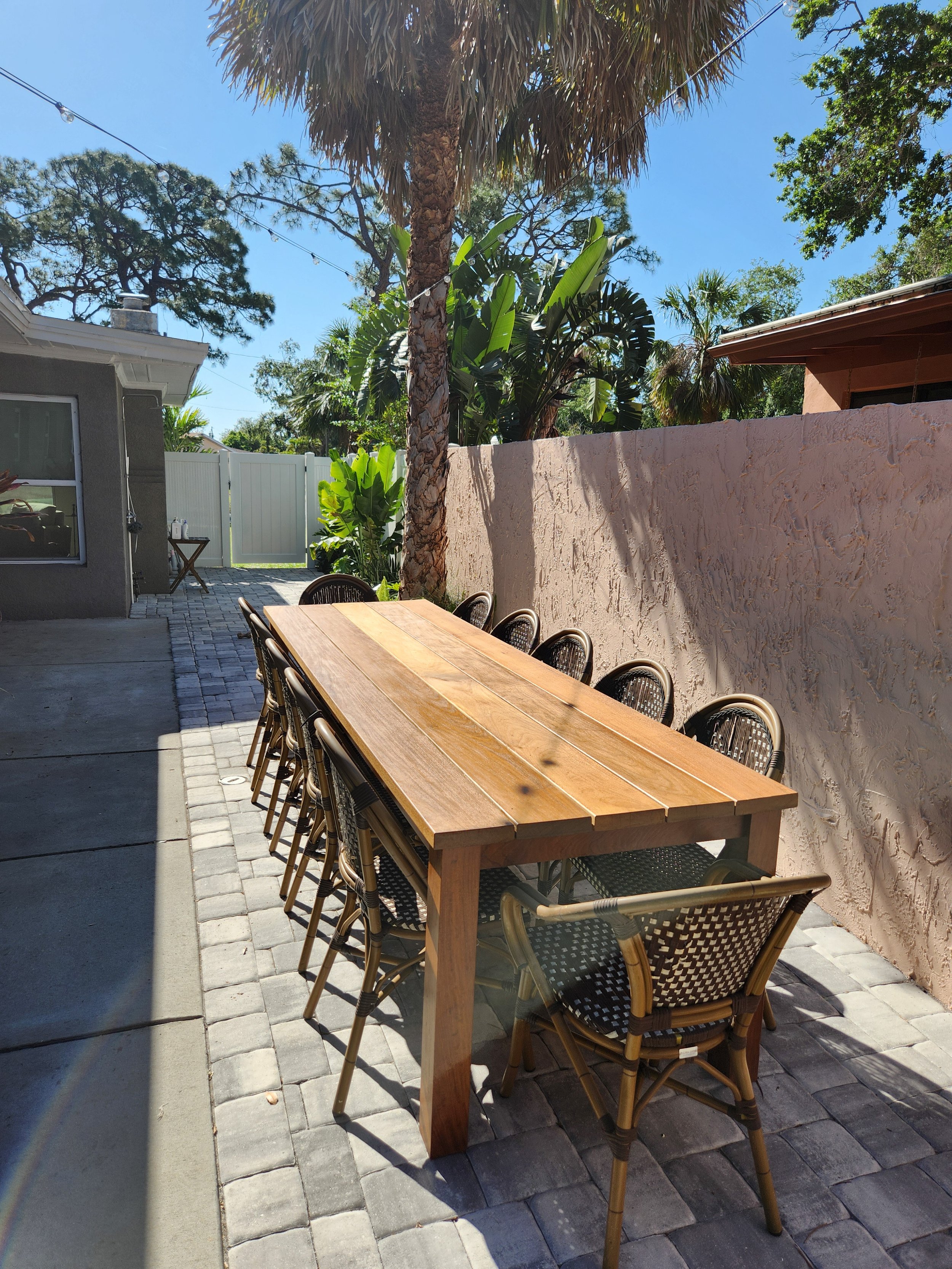 Wooden outdoor dining table with ten chairs on a paved patio, surrounded by palm and other trees, with a textured beige wall and a brown building on the right, a gray house with a window on the left, and a white fence with a gate in the background under a bright blue sky.