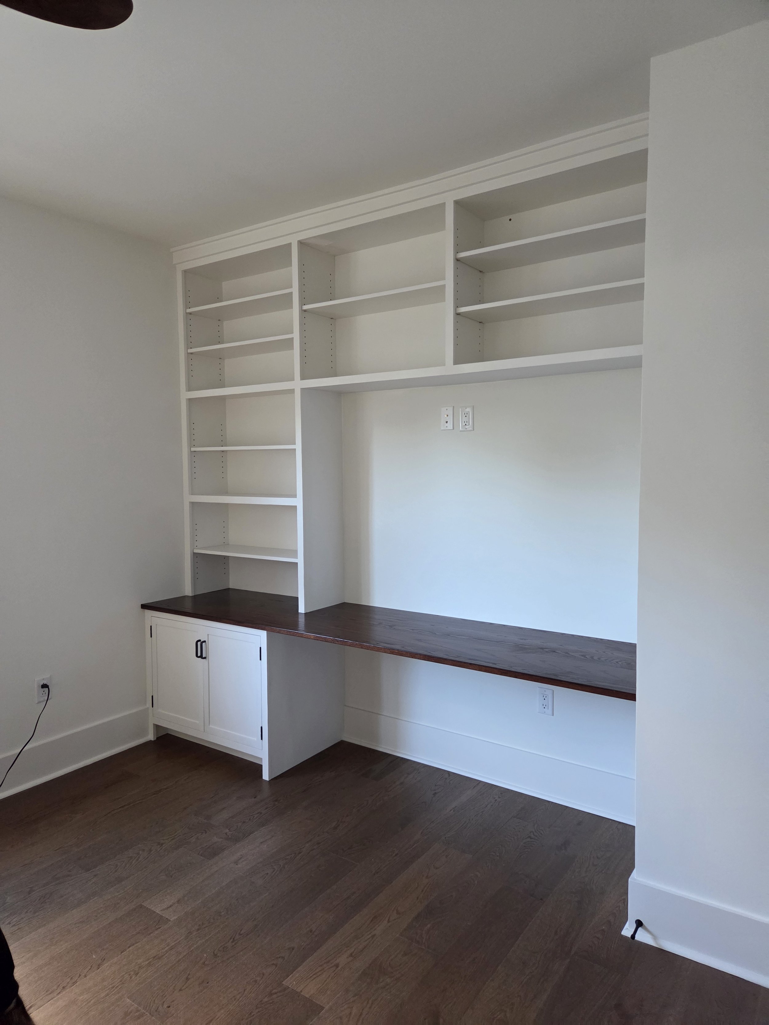 Empty built-in white bookshelf with multiple shelves mounted on a white wall in a room with dark hardwood floor.