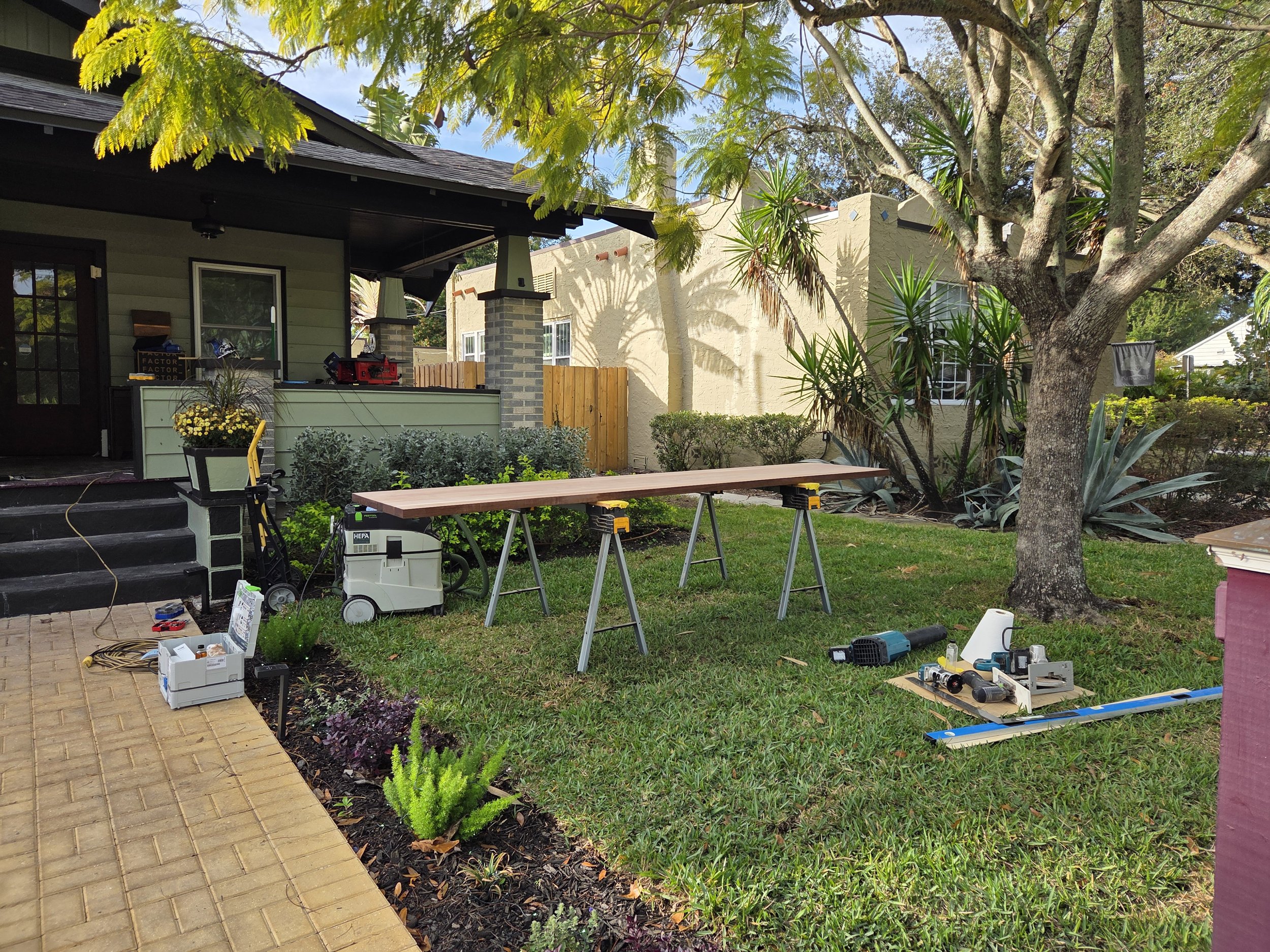 A backyard yard under construction with tools and materials on the grass and a work table. There is a large tree, a house with a porch, and a beige wall in the background.