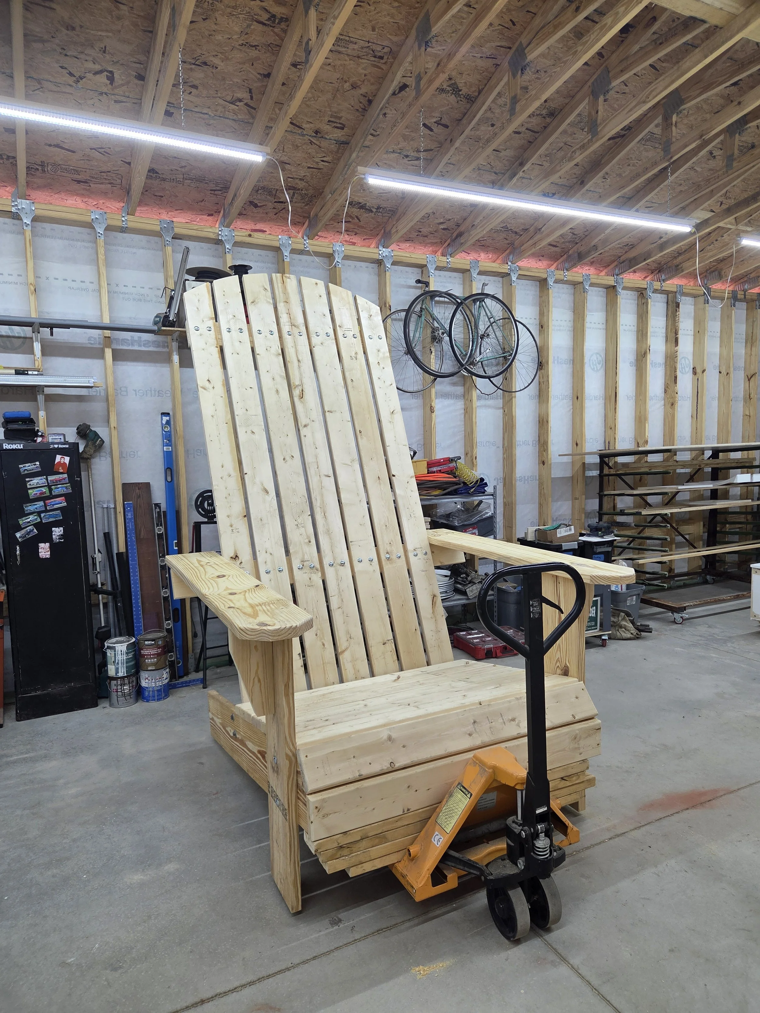 A wooden outdoor bench with a backrest and armrests, stationed in a workshop or garage with unfinished walls, hanging bikes, tools, and construction materials.