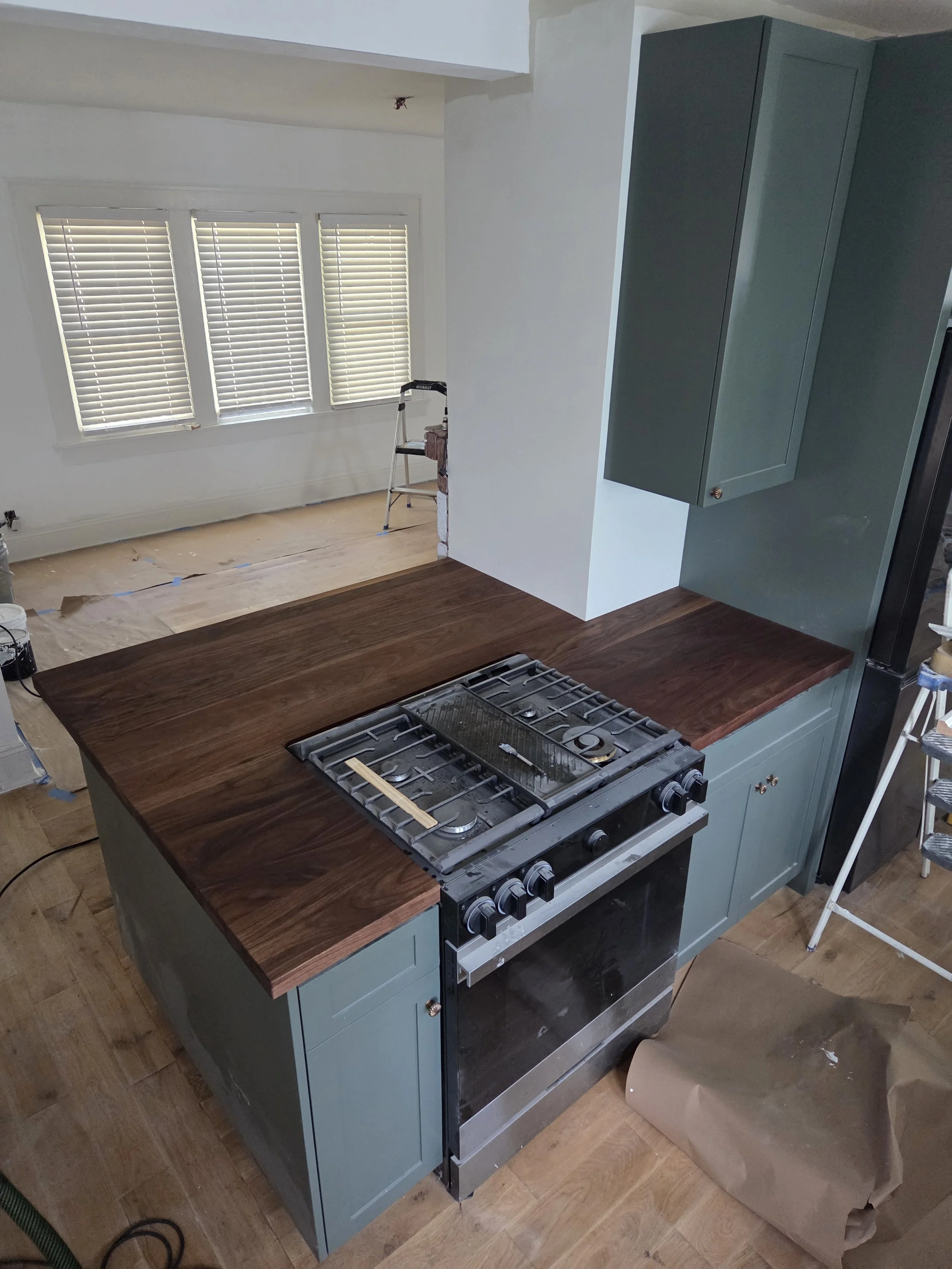 View of a kitchen under renovation with a stove, green cabinetry, a wooden countertop, and a window with blinds.