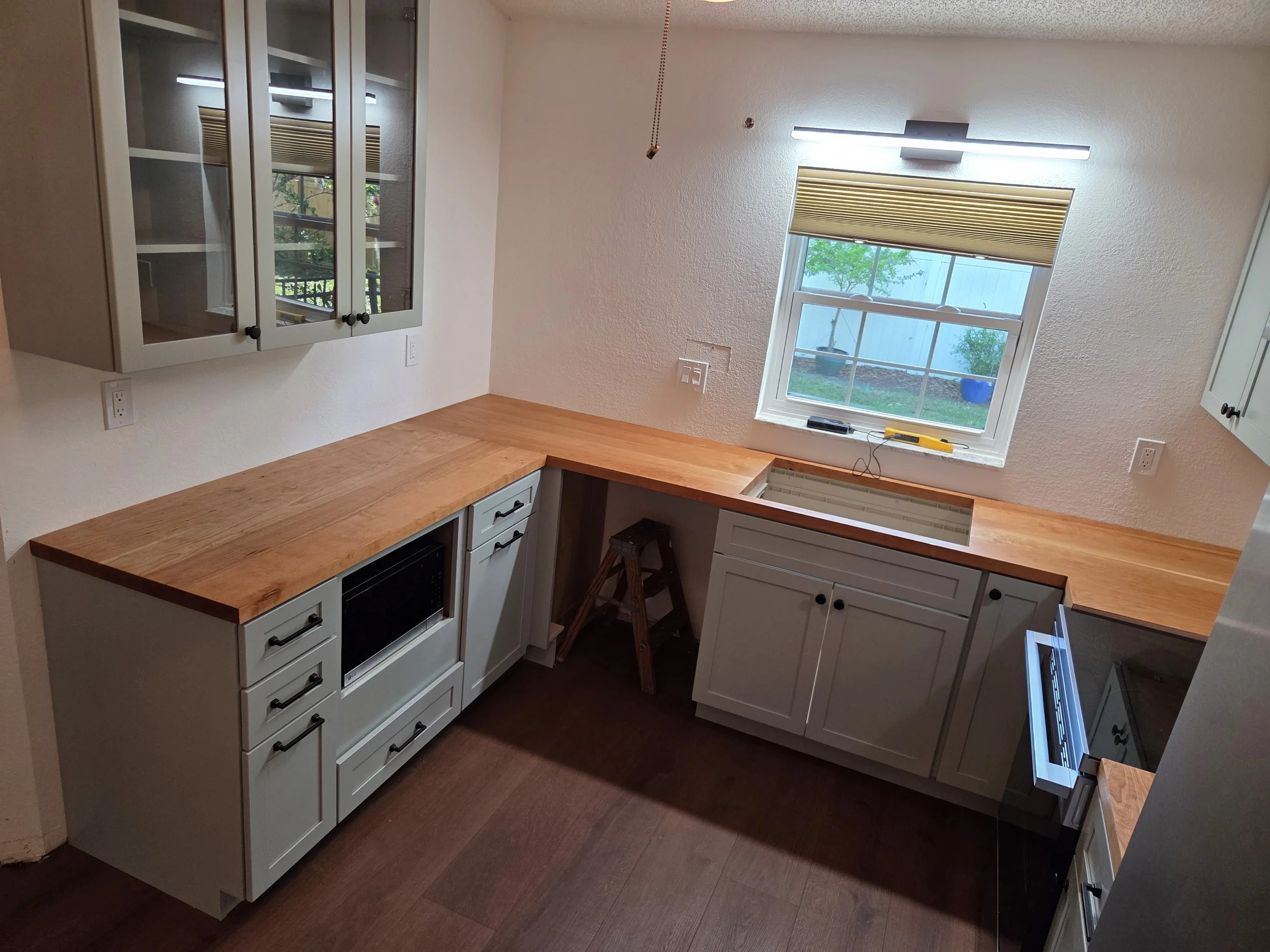A kitchen with white cabinets, a wooden countertop, a window with a beige blind, and a ceiling light. There are power outlets on the wall and a small step near the window.