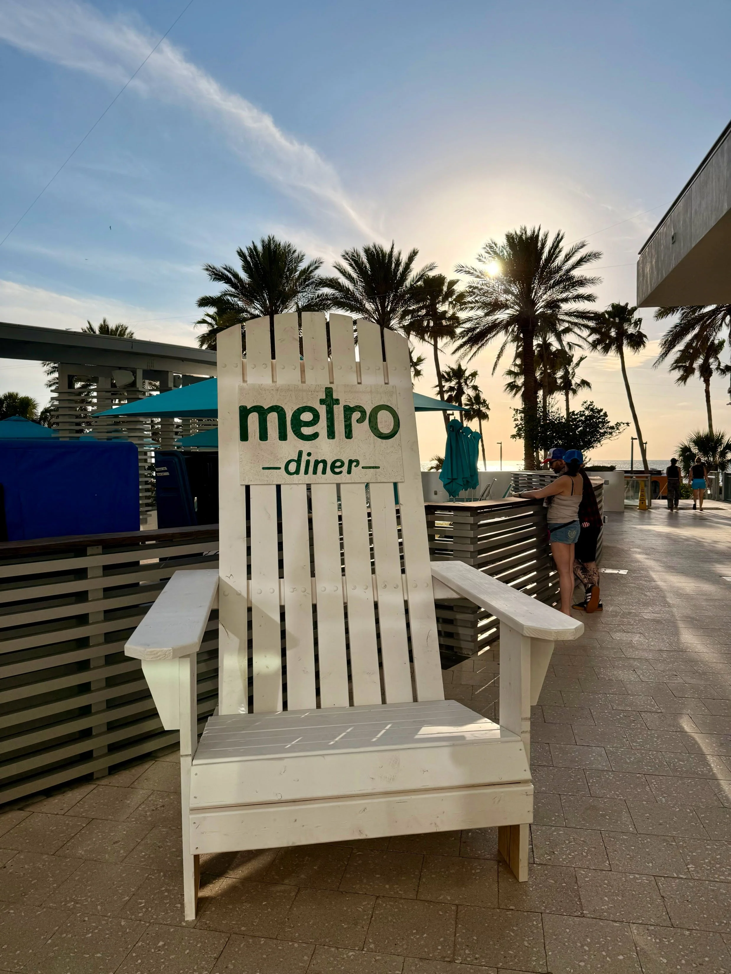 Empty white wooden chair in front of a Metro Diner sign with palm trees and sunset in the background.