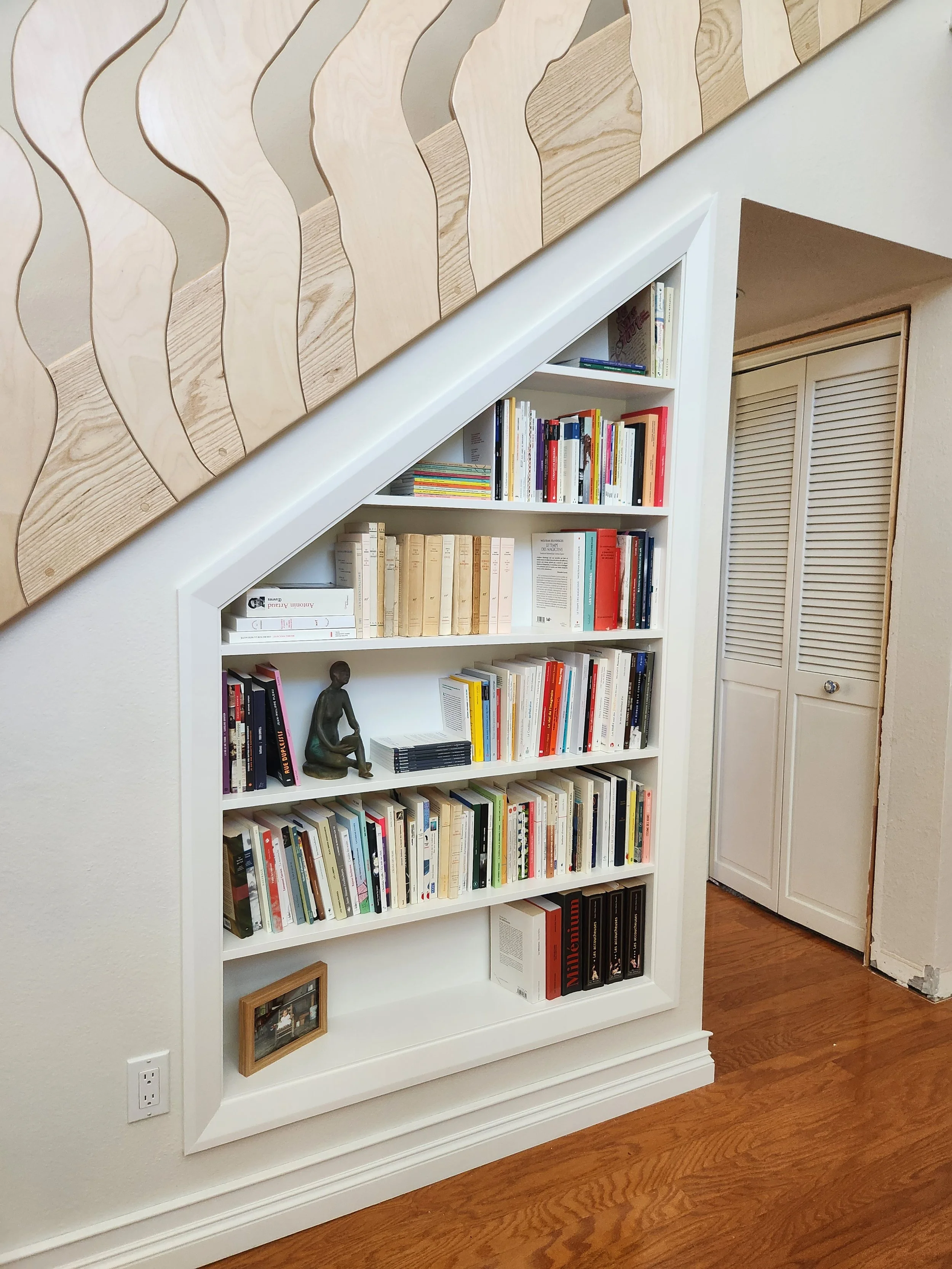A white bookshelf filled with books, a small sculpture of a seated person, and a framed photograph. The bookshelf is positioned against a white wall in a room with wooden flooring and a closed, louvered closet door.
