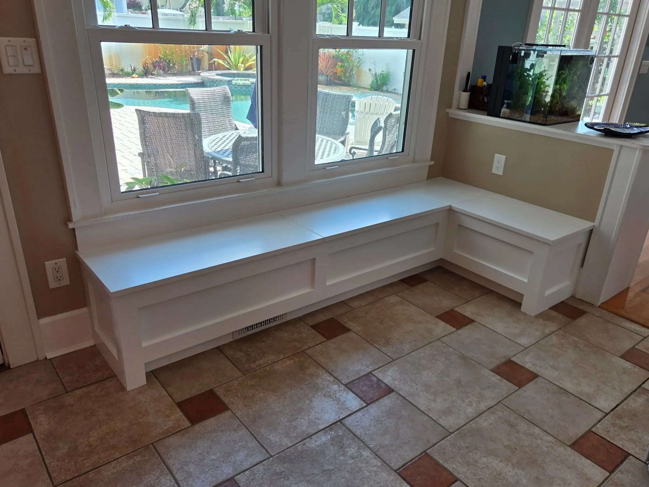 View of a large window overlooking a backyard with a swimming pool, patio furniture, and surrounding greenery. Inside, there is a built-in white bench beneath the window and a small aquarium on a corner shelf.