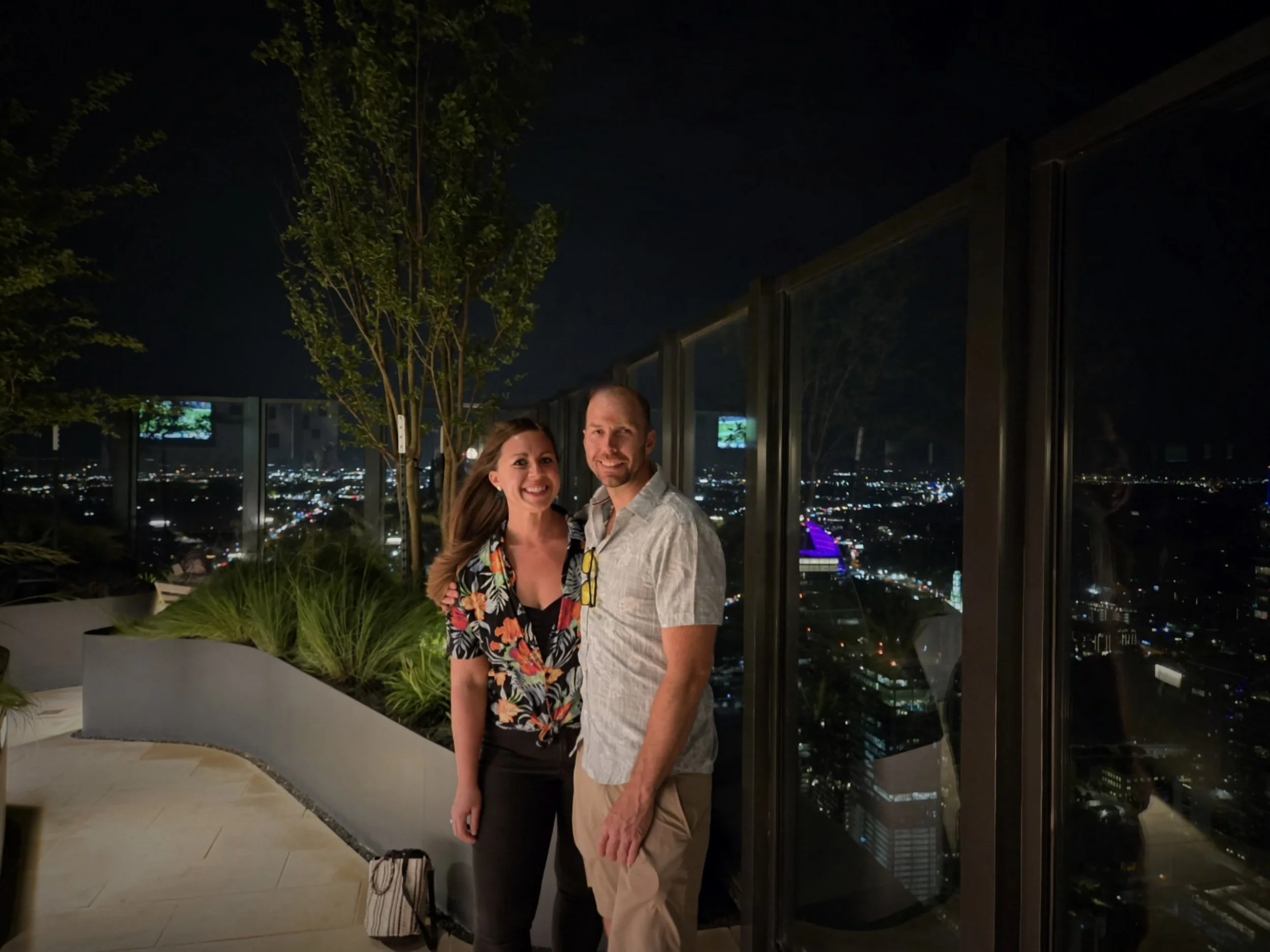 A smiling couple standing close together on a balcony at night, with city lights and tall buildings in the background, overlooking a cityscape.