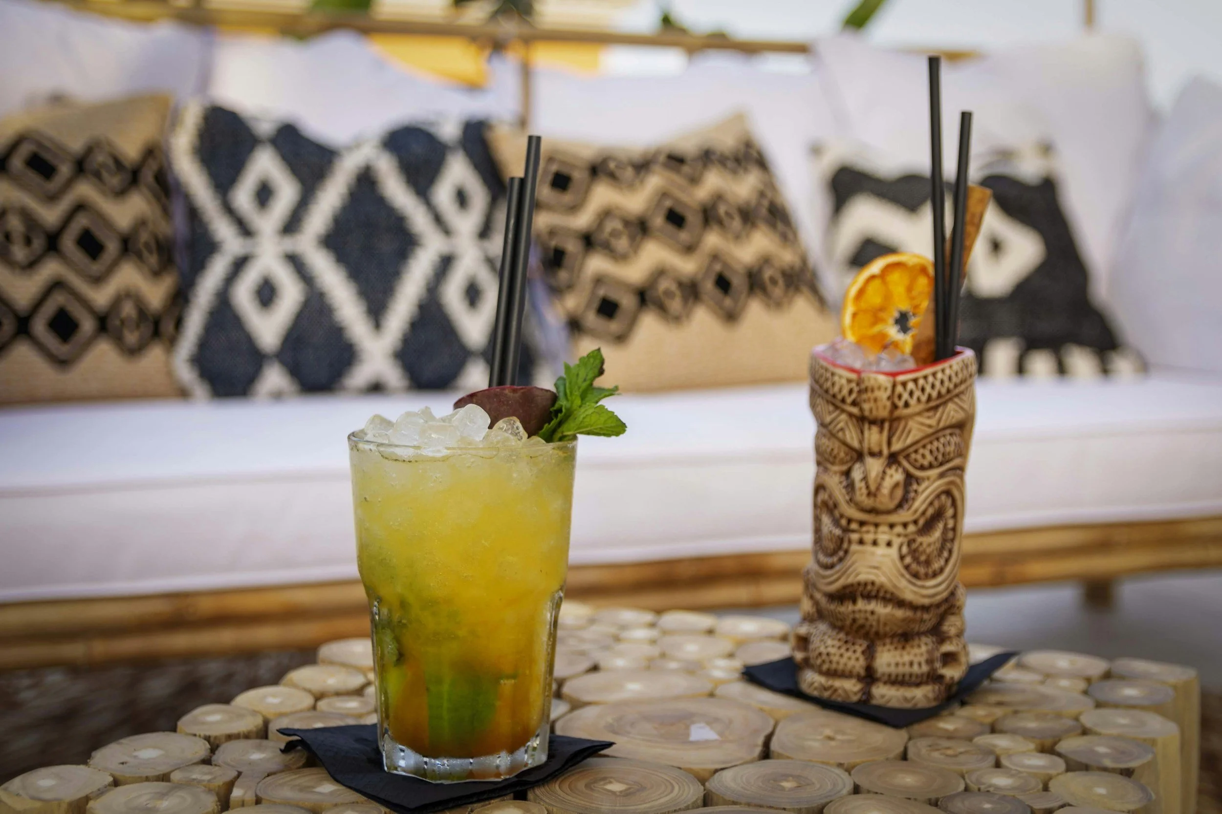 A tropical cocktail garnished with a slice of orange and mint leaves on a wooden table, and a tiki-style drink holder with two black straws, in front of a background with cushions and a patio umbrella.