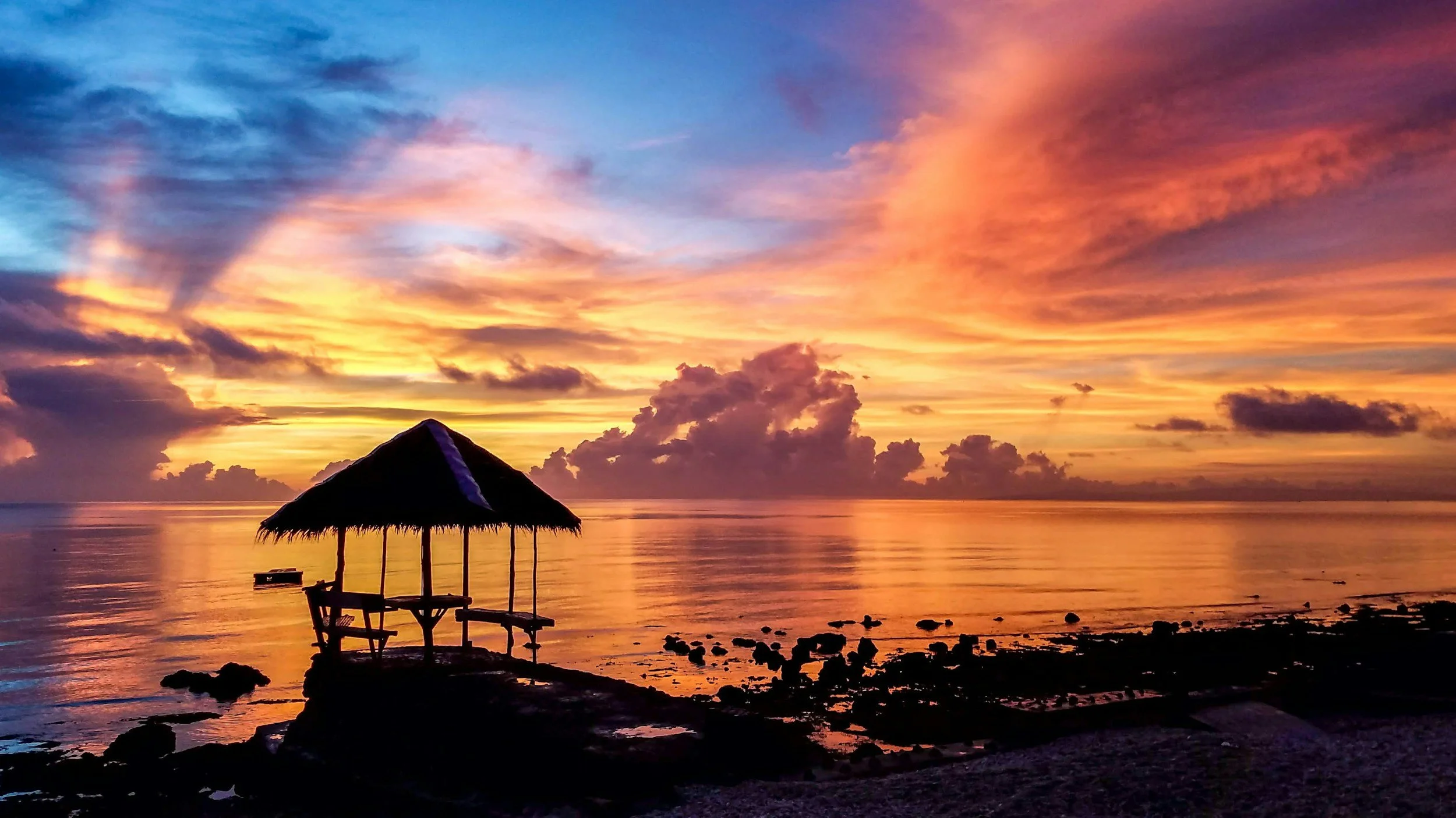 Sunset over the ocean with colorful clouds in pink, orange, and purple hues, and a thatched-roof tiki hut on the beach in the foreground.