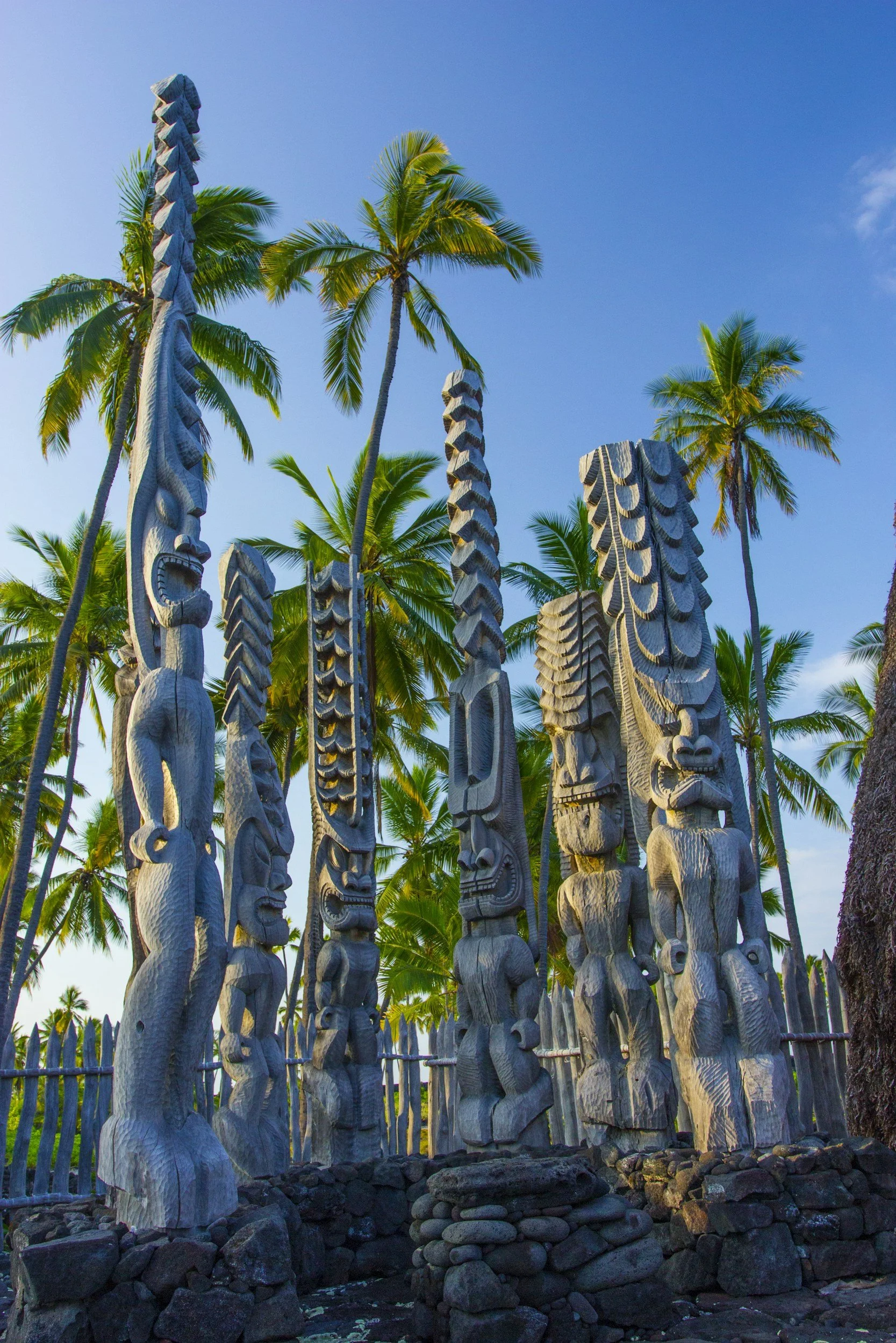Wooden Maori statues standing among green palm trees against a blue sky.