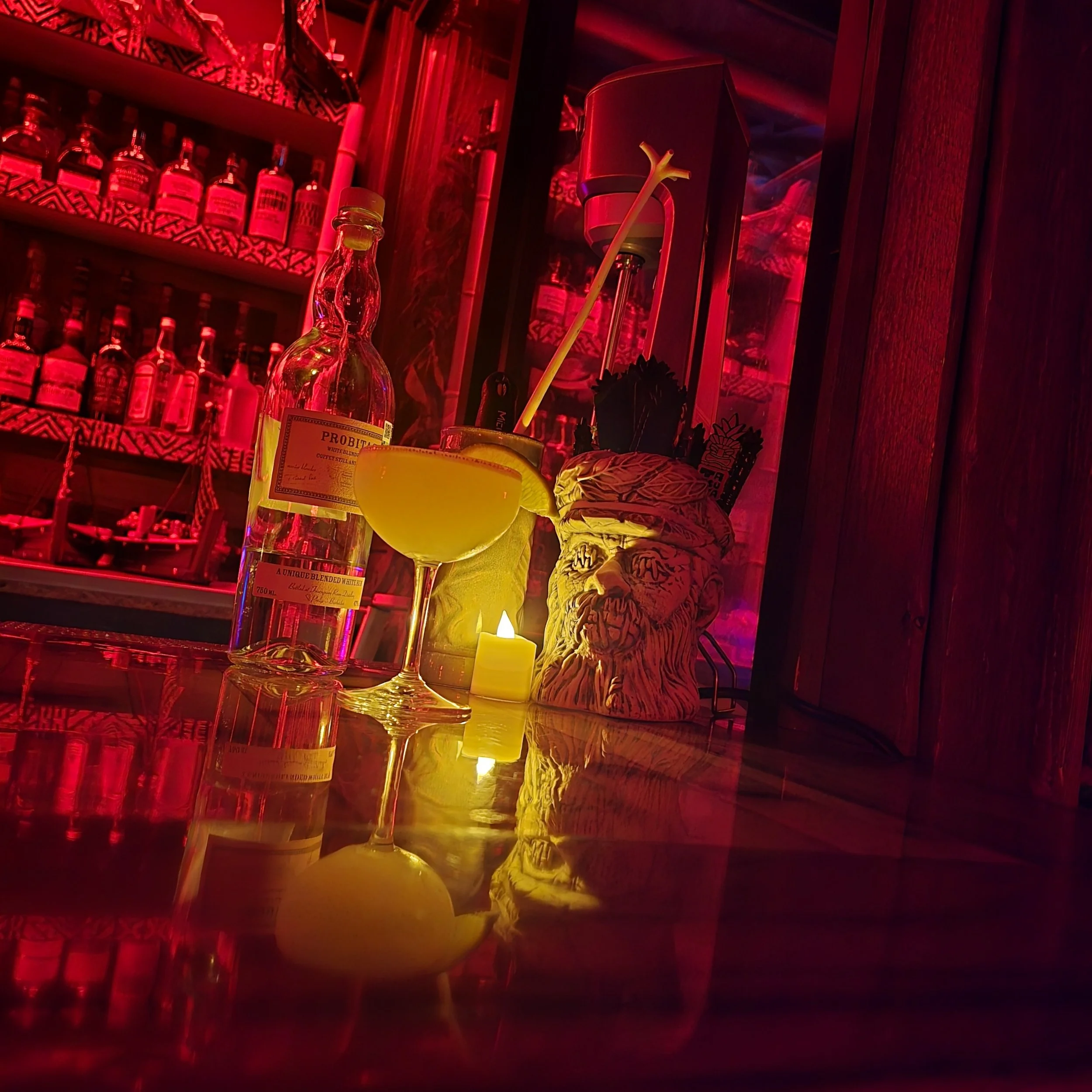 A dimly lit bar or restaurant with red lighting, shelves with bottles and decorative items, a candle, and some glassware on a reflective surface.