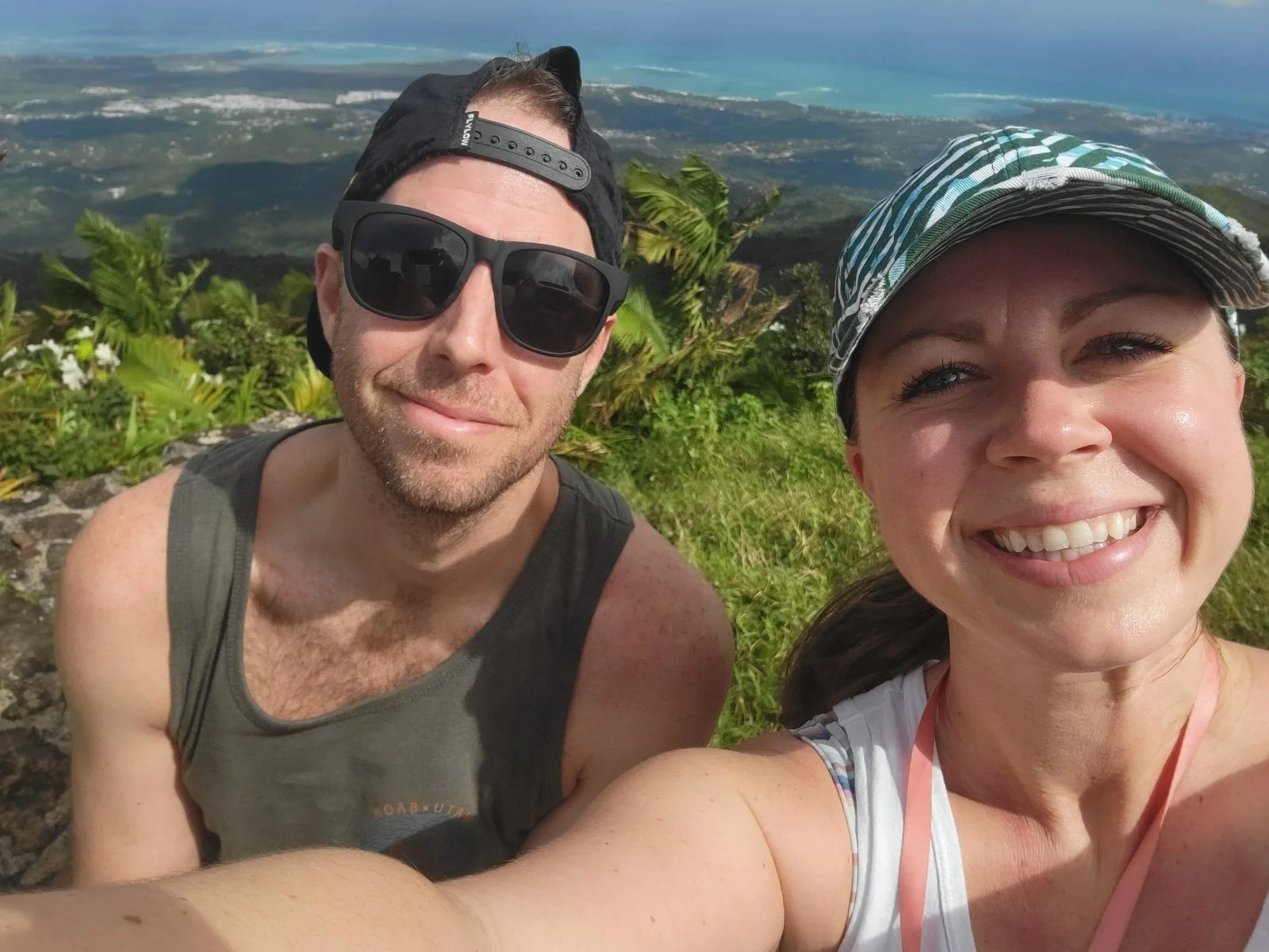 A man and woman taking a selfie outdoors with lush green vegetation and a distant view of the coastline in the background, both smiling and wearing casual summer clothing and hats.