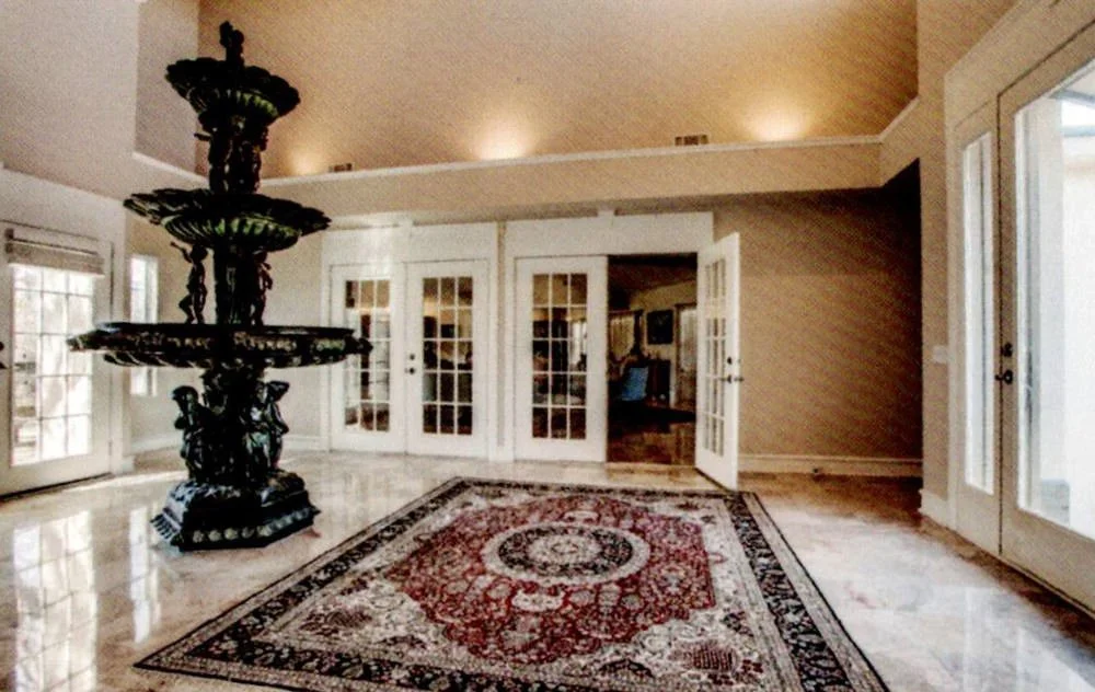 Large, elegant living room with glass-paned French doors, a vintage multi-tiered fountain, a decorative area rug, and hardwood floors.