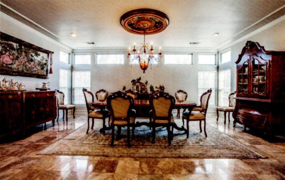 A dining room with a wooden oval table surrounded by seven upholstered chairs, a chandelier hanging above, large windows on three sides, a sideboard on the left, and a large wooden china cabinet on the right, with a decorative rug on the marble floor
