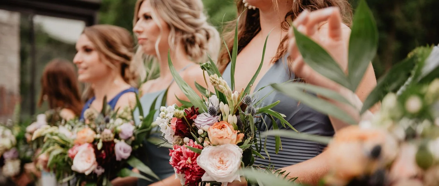 A line of women holding bouquets of mixed flowers, wearing dresses, at an outdoor event.
