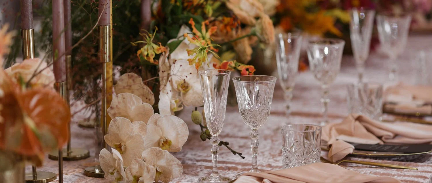 Elegant table with crystal glassware, peach-colored napkins, and floral centerpieces with orchids and autumn-colored flowers.