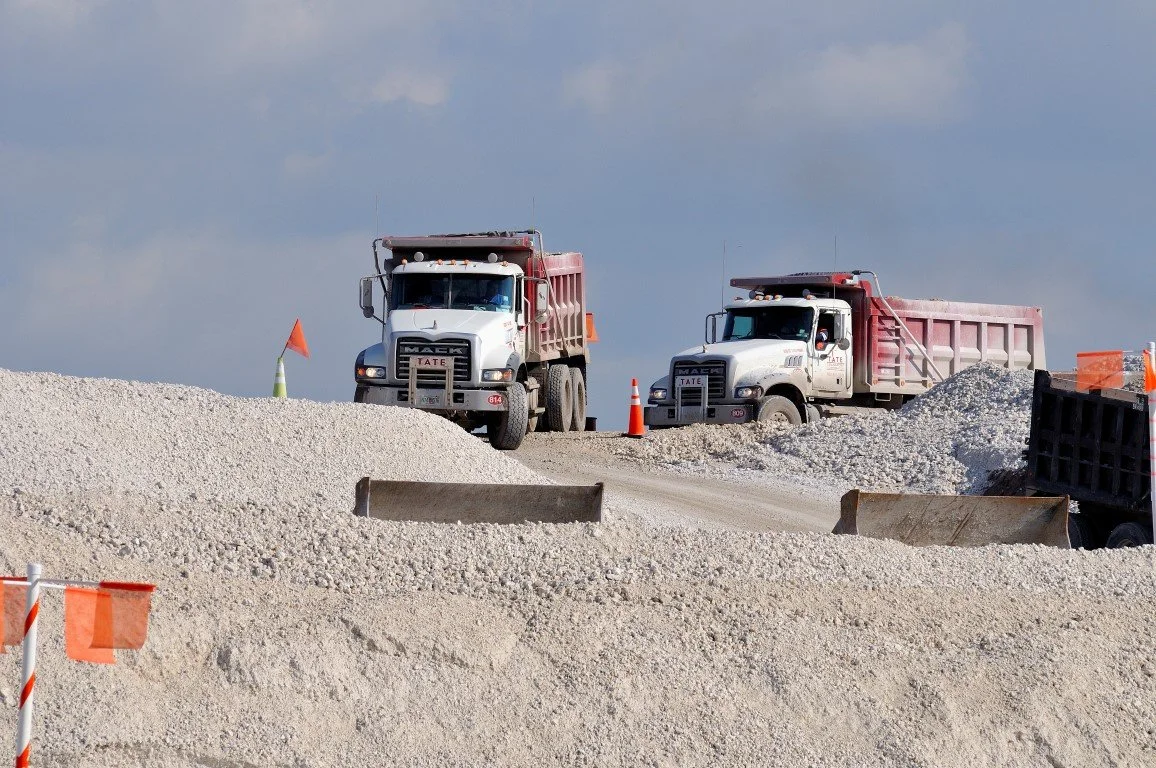 Construction site with two large dump trucks on a gravel road, multiple orange safety flags and cones visible.