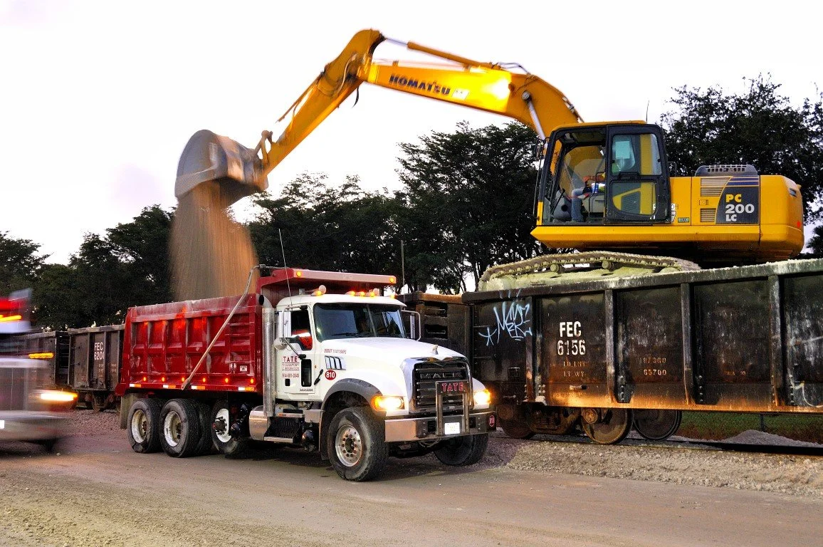 A yellow excavator deposits dirt into a red dump truck while a black train car is adjacent on tracks; the scene appears to be construction or railway maintenance work.