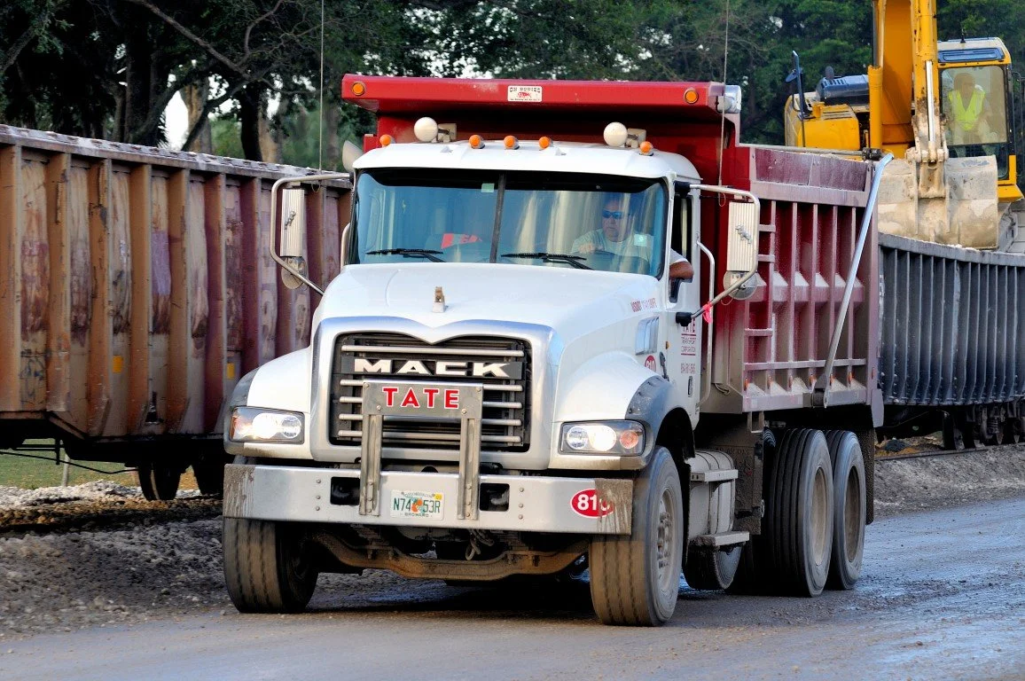 A white Mack dump truck with the name Tate on the front, driving on a construction site with dirt and other trucks in the background.