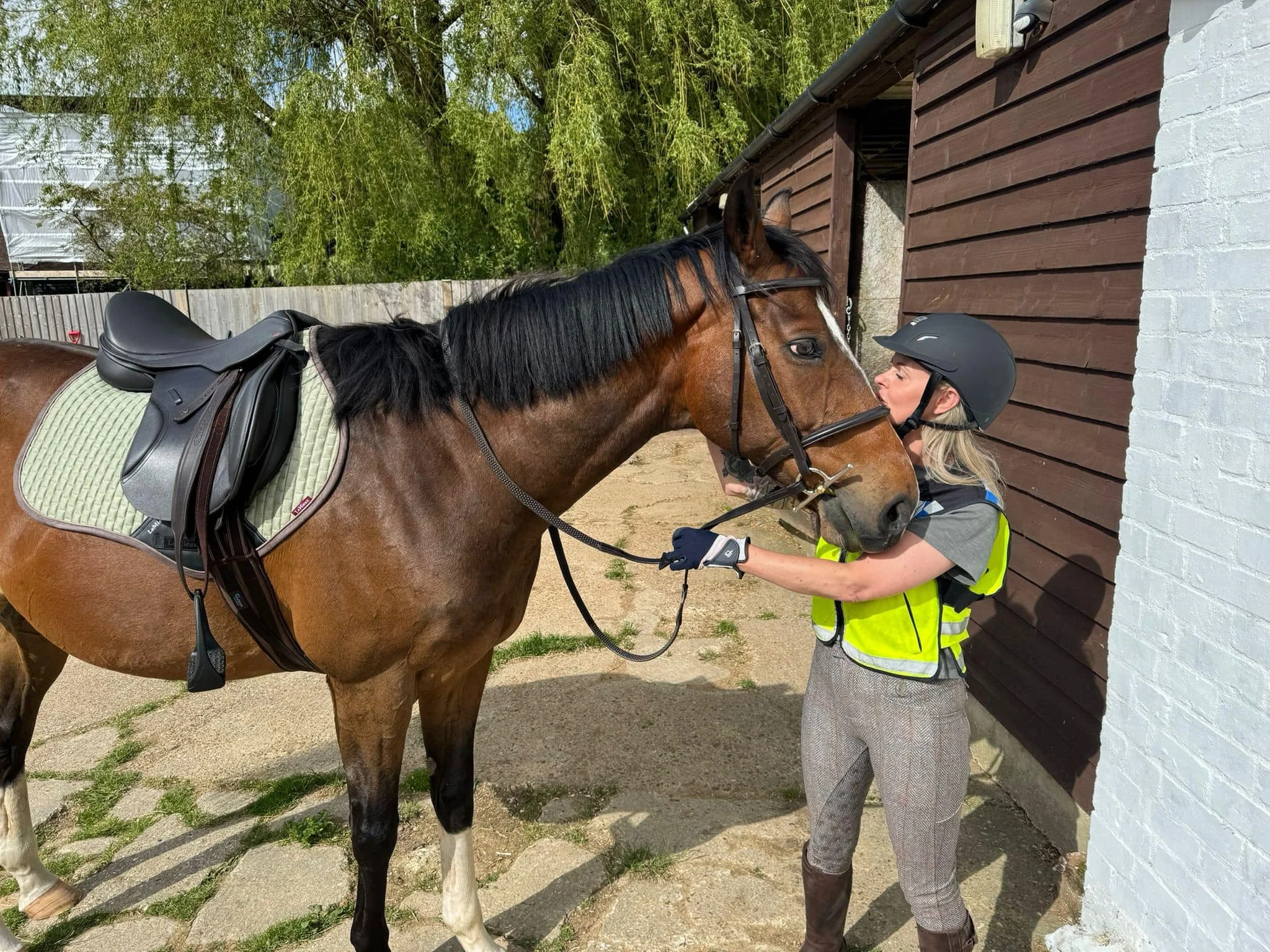 A woman in riding gear, including a helmet and yellow safety vest, kisses a brown horse with a black mane outside a stable.
