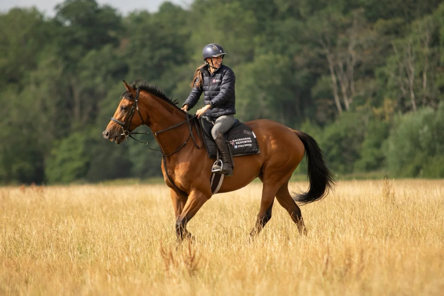 A woman riding a brown horse across a grassy field with trees in the background.