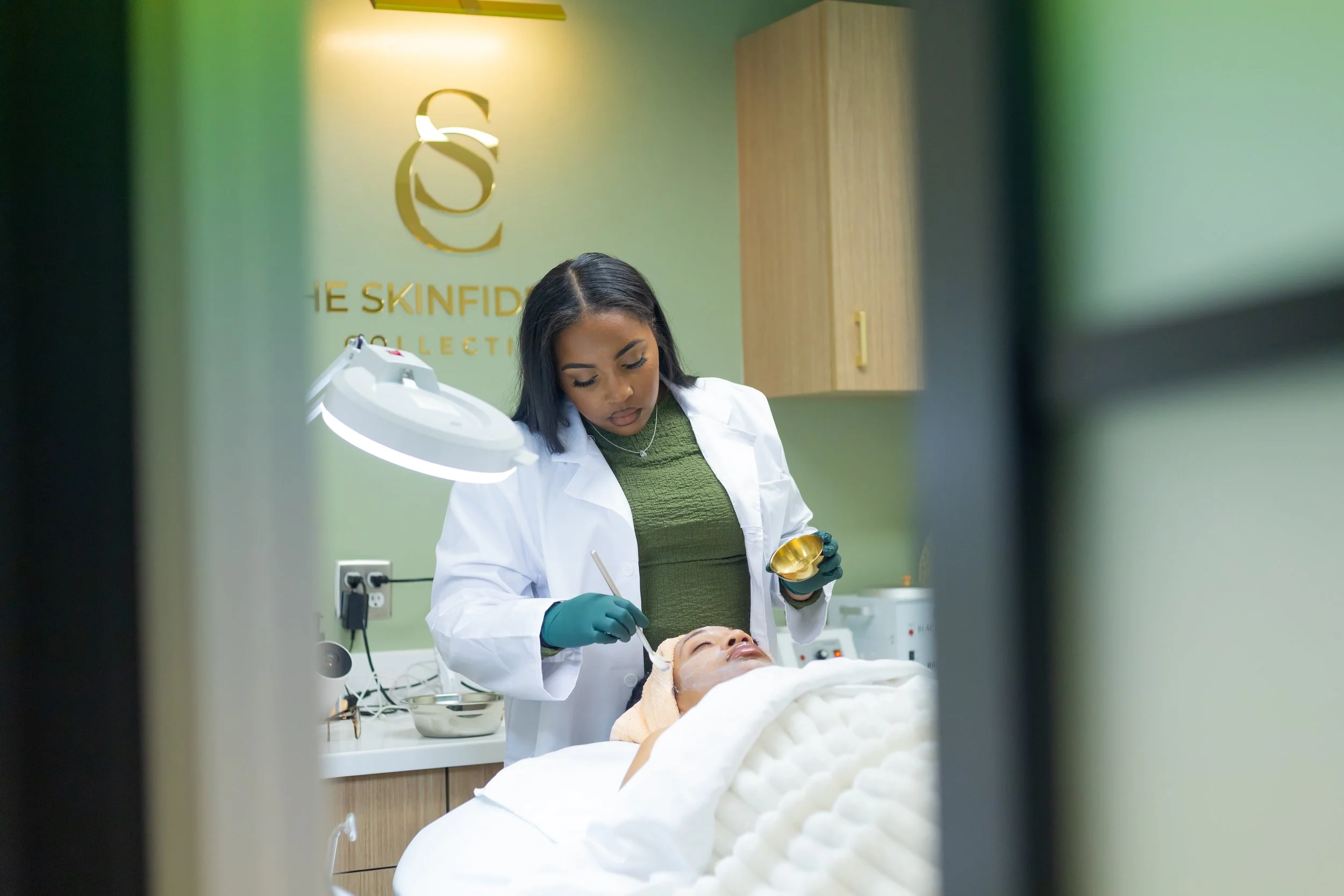 A woman lying on a treatment bed with her eyes closed, receiving a facial treatment from a skincare professional in a clinic with green and wooden decor. Skinfidence Collective.