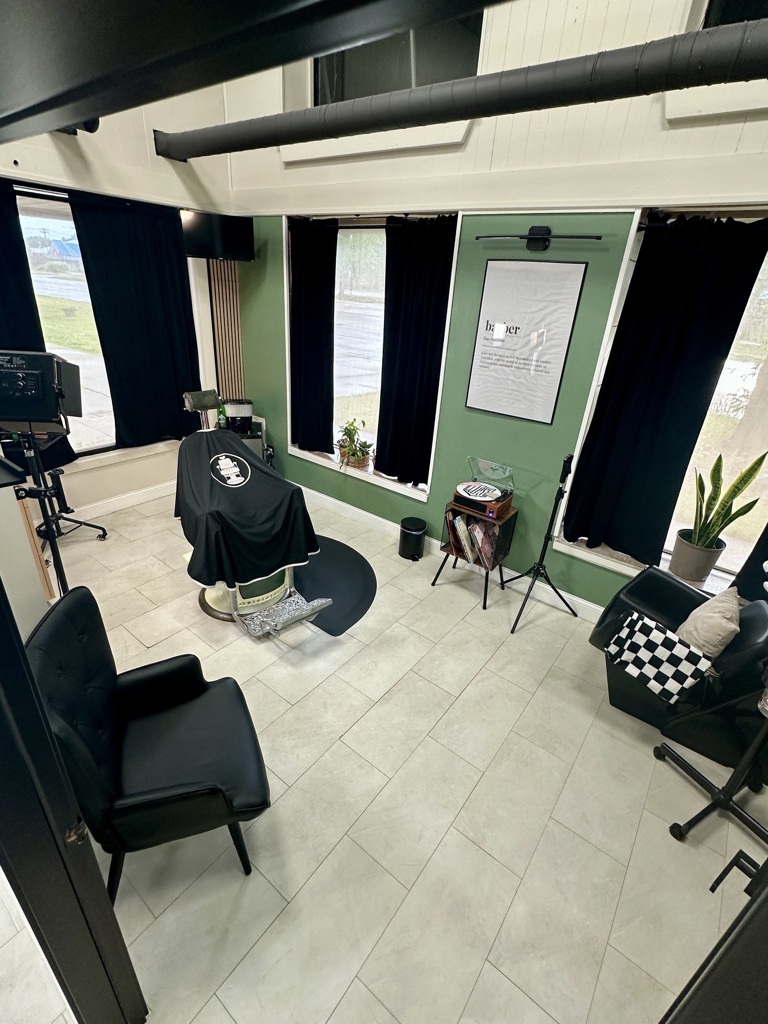 A room with black chairs, a green wall with a window, black curtains, a plant, and music equipment, viewed from a door's perspective. Barbers and Brethren barber shoppe.