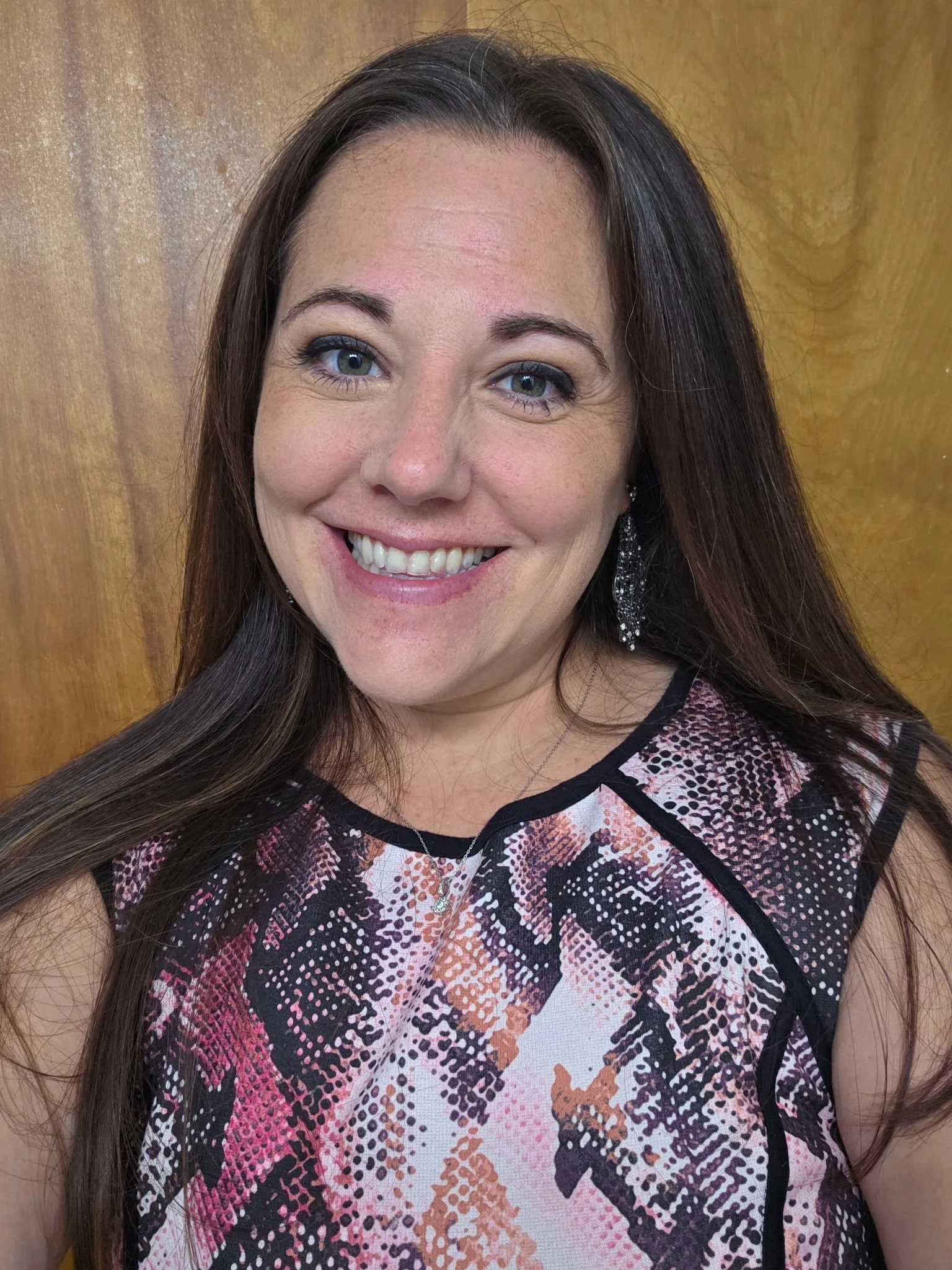 Close-up of a woman smiling, with long brown hair, wearing a patterned top with black, pink, and orange colors, and a silver necklace with a small pendant, standing against a wooden background. Jessica Kathleen's Hair co.