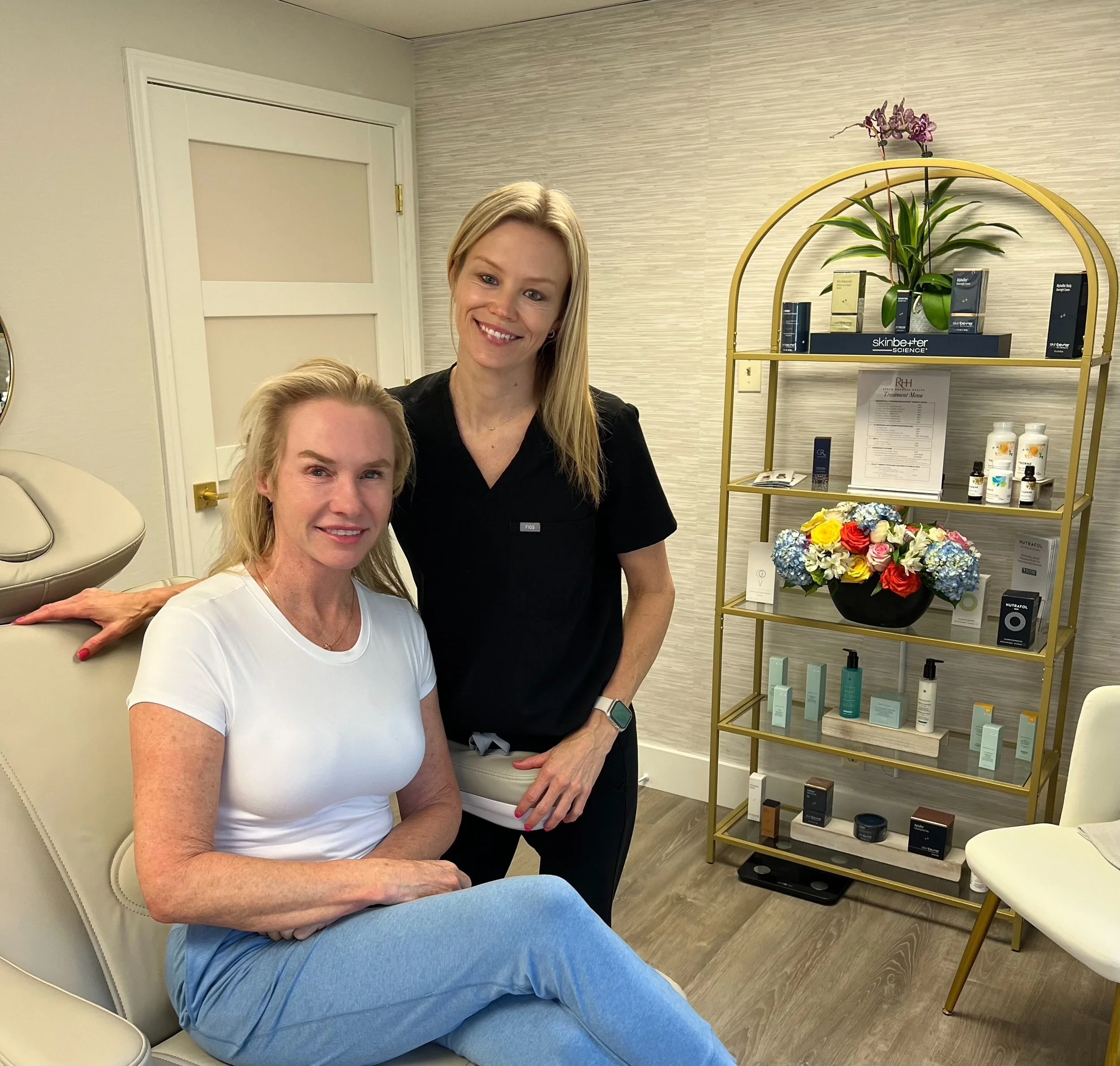 A woman with blonde hair on a beige chair, smiling and talking about peptide therapy with a woman with blonde hair standing behind her, smiling, in a skincare or beauty clinic with a gold shelf displaying skincare products.