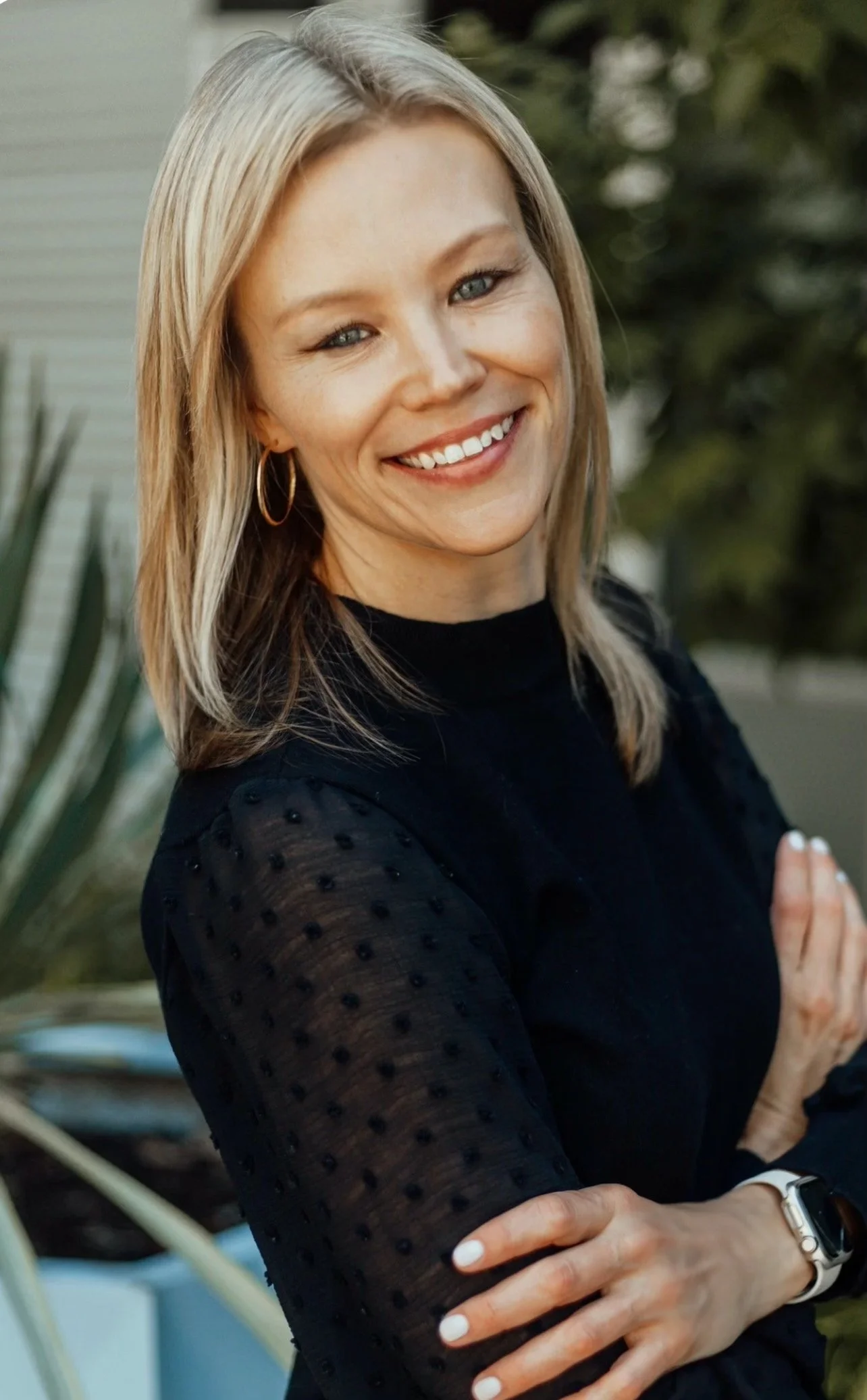 A woman with blonde hair and blue eyes smiling, wearing a black top with sheer, dotted sleeves, and jewelry including hoop earrings and a watch, standing outdoors with blurred greenery in the background.
