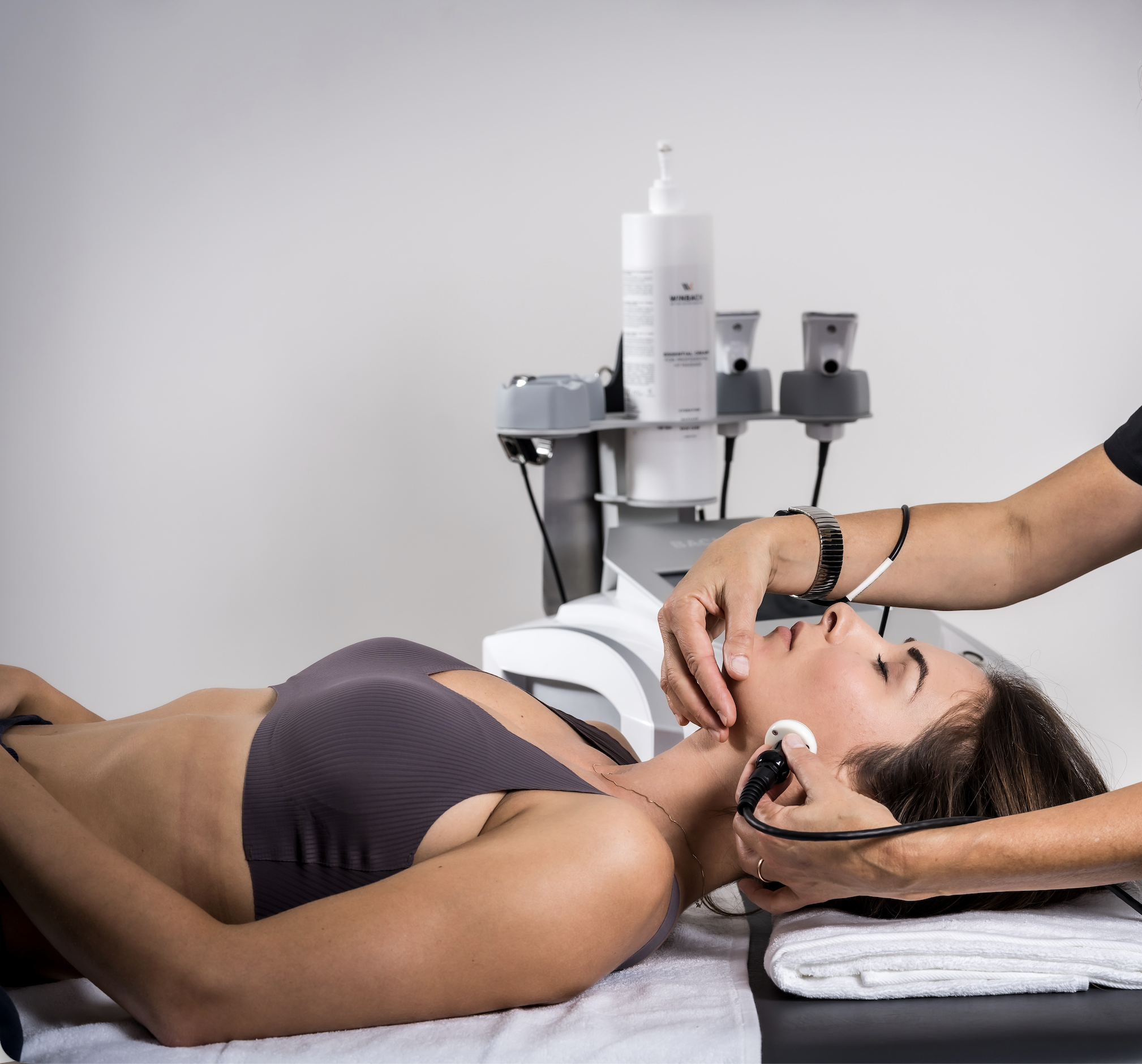 A woman lying on a medical table receiving a facial treatment with a skincare device, while a healthcare professional administers the treatment using a handheld device near the woman's face in a clinical setting.
