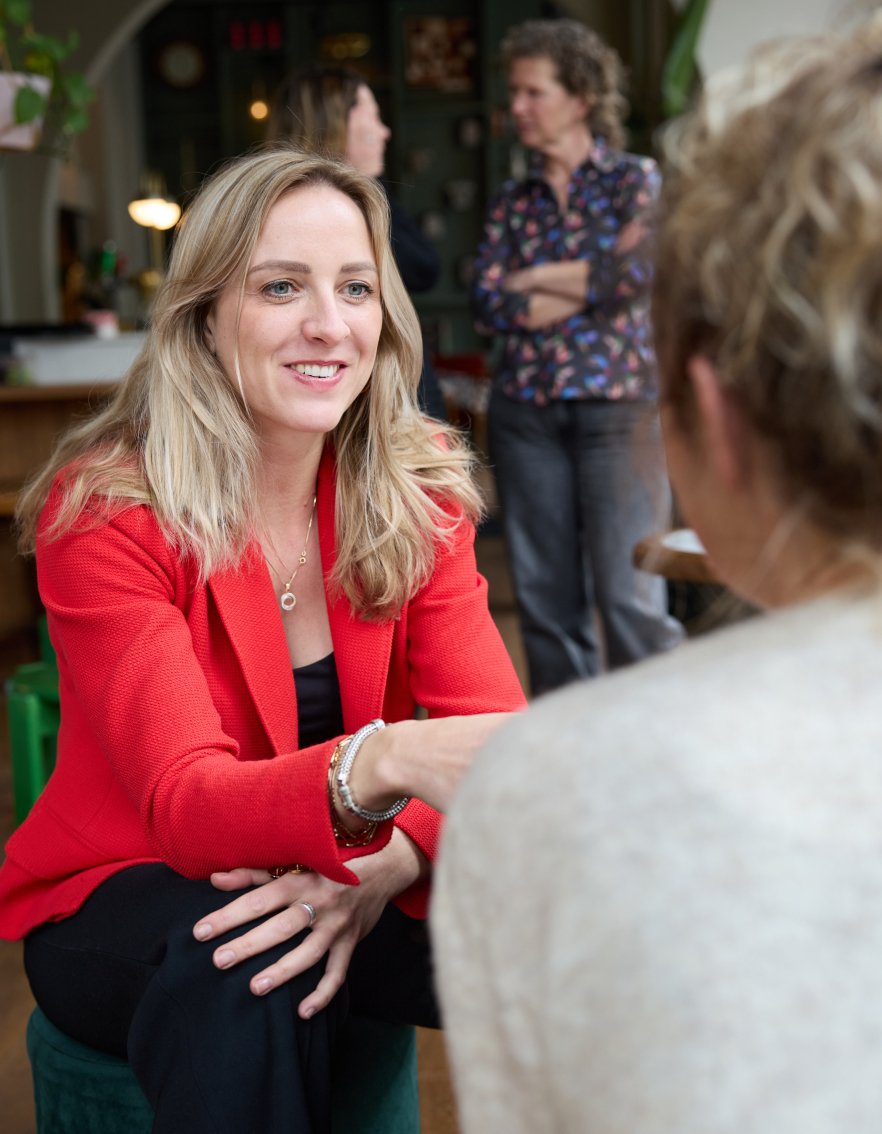 Een vrouw in een rode jas glimlacht terwijl ze met een andere persoon praat, terwijl op de achtergrond twee vrouwen in gesprek zijn in een gezellige binnenruimte.