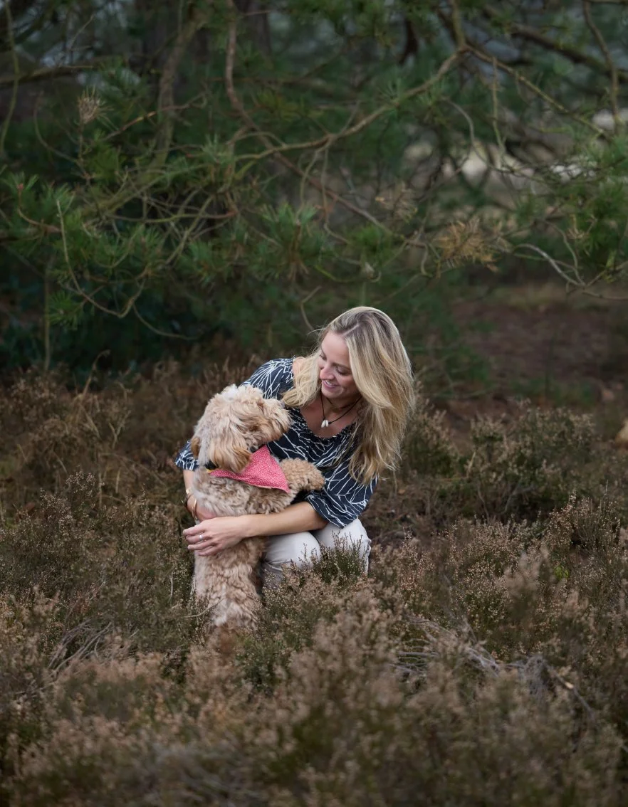 Vrouw met blond haar die knuffelt met een lichtbruin, klein hondje met een roze bandana in een bosrijke omgeving.